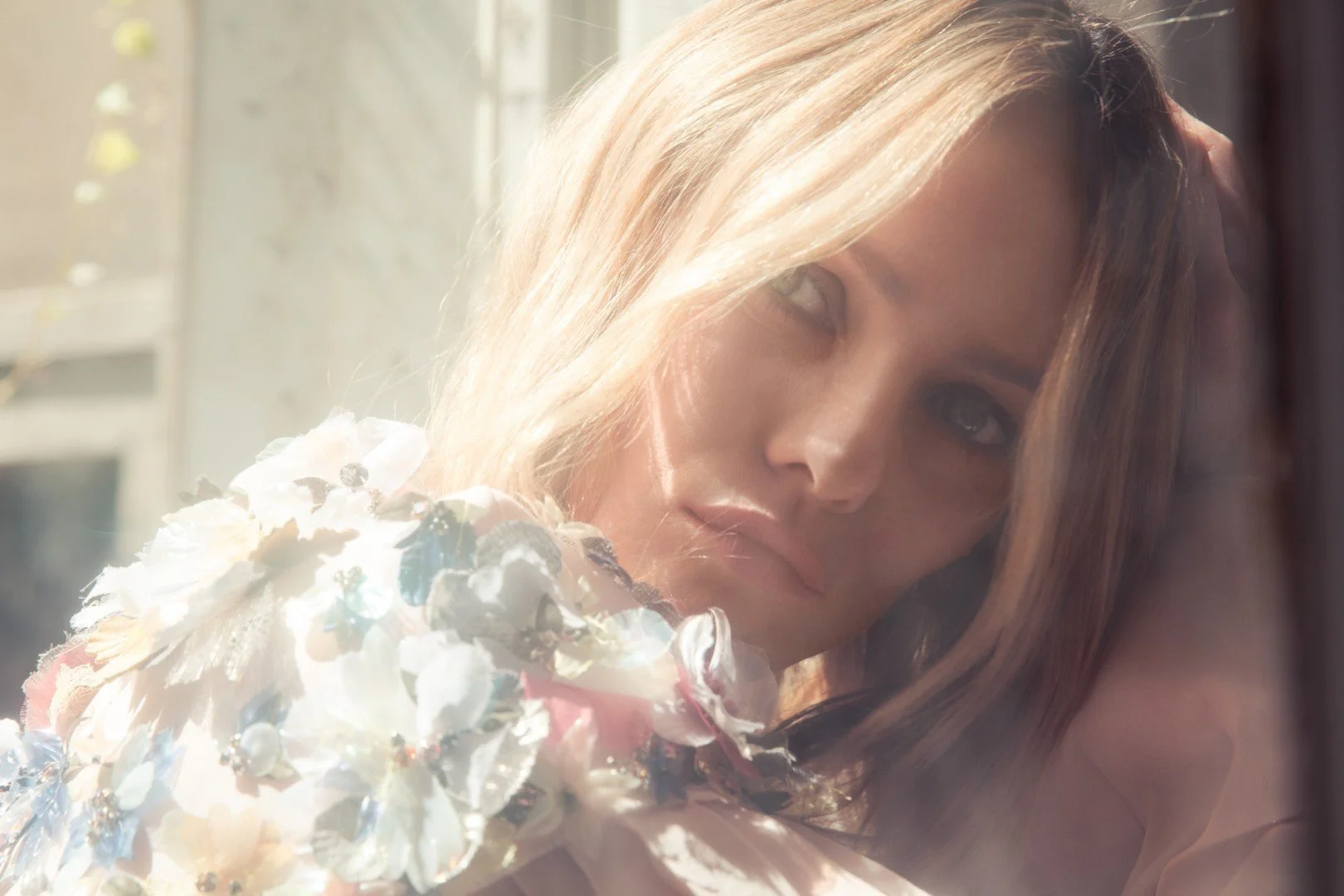 A young woman with blonde hair and blue eyes leans against a window, looking directly at the camera. She is holding a bouquet of white and pastel-colored flowers, with soft sunlight illuminating her face.