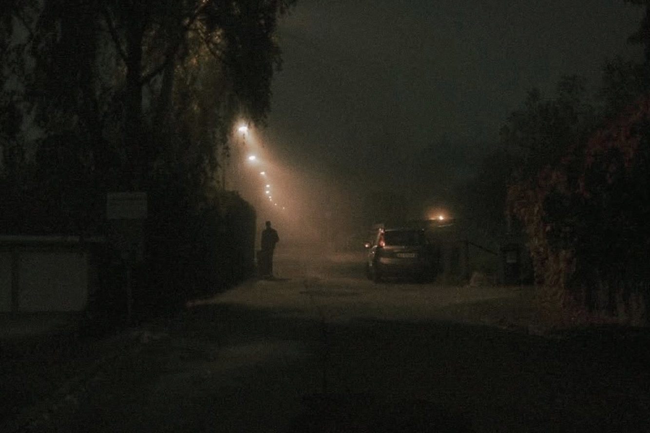 A dark, foggy street at night with a person walking and parked cars along the side, illuminated by streetlights