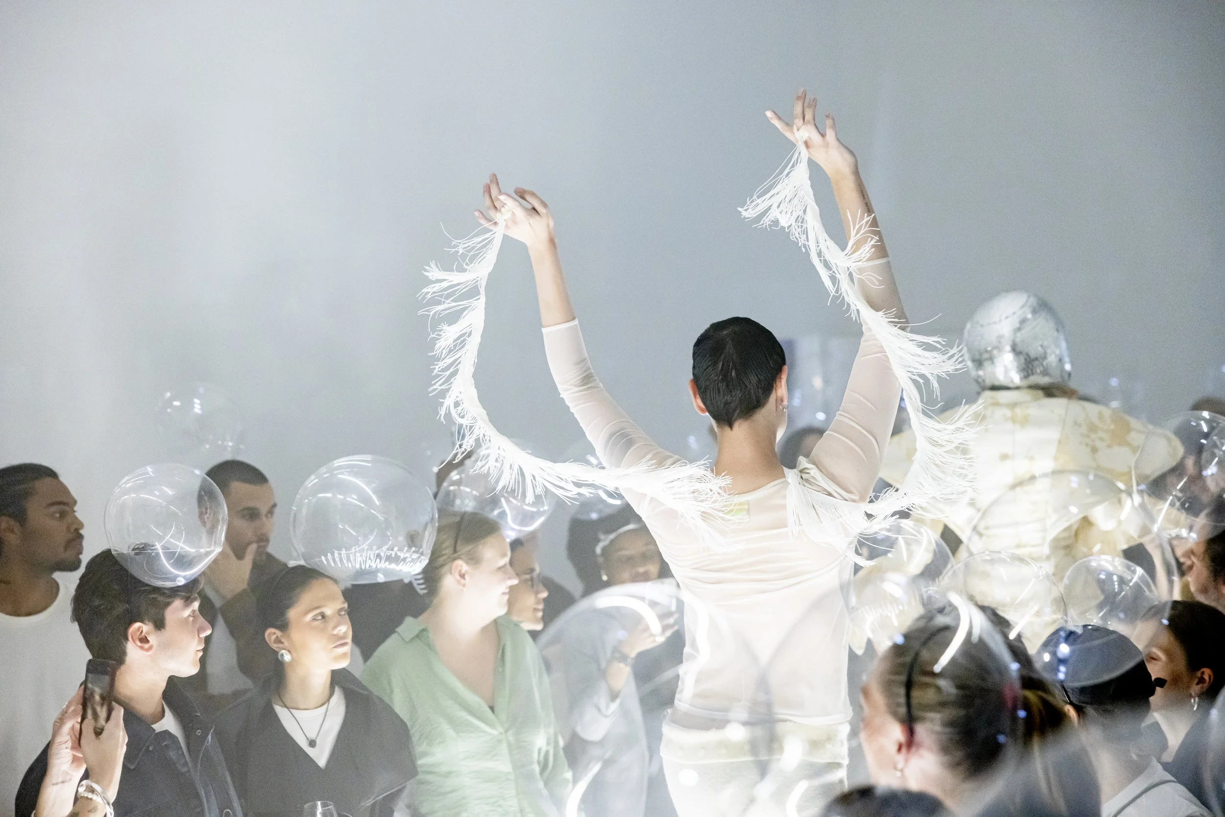 A performer with short black hair wearing a white fringe costume is dancing with arms raised in front of a crowd at an event surrounded by transparent balloons.