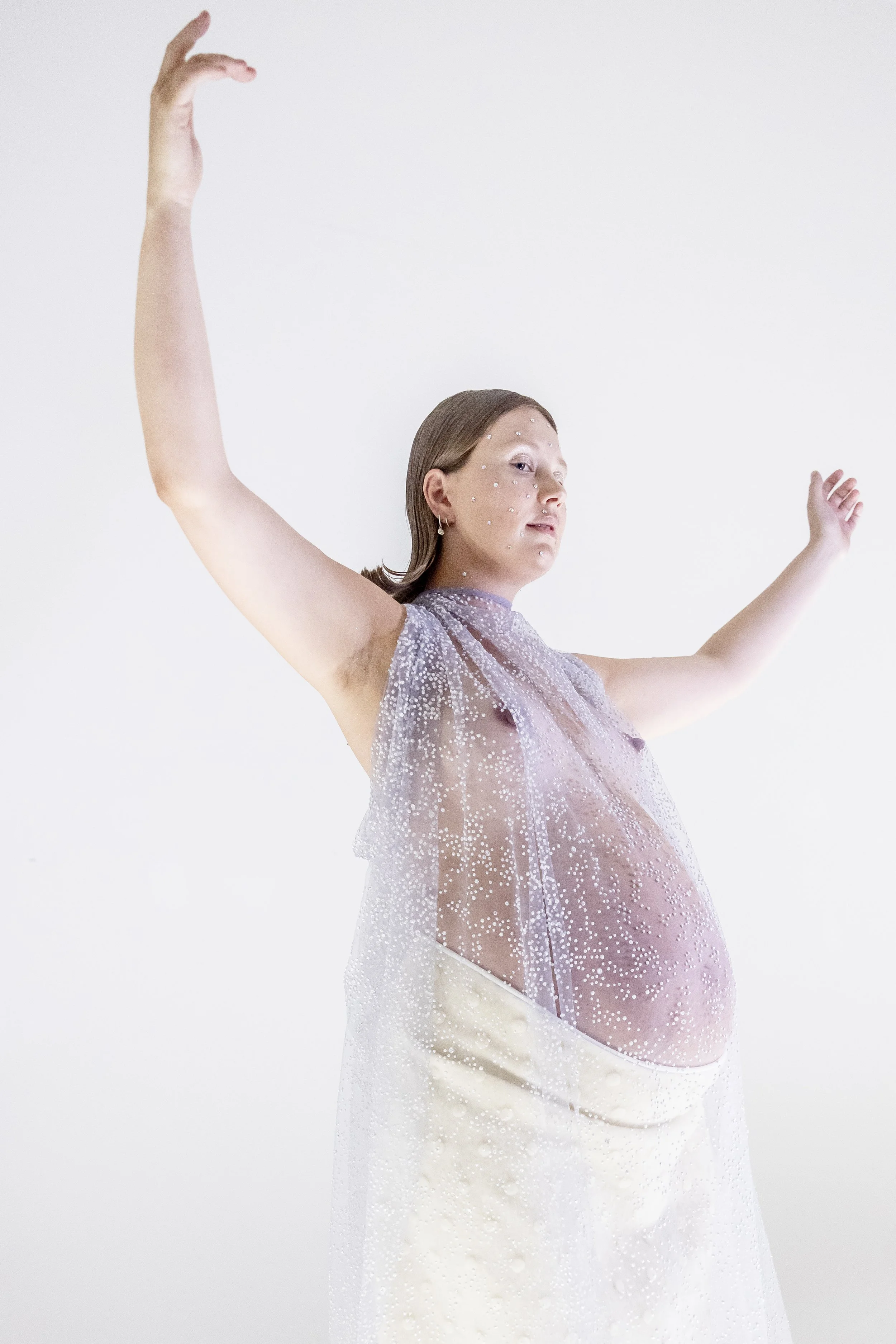 A pregnant woman with light skin, wearing a sheer, sparkly fabric draped over her shoulders, poses with her arms raised against a plain white background.