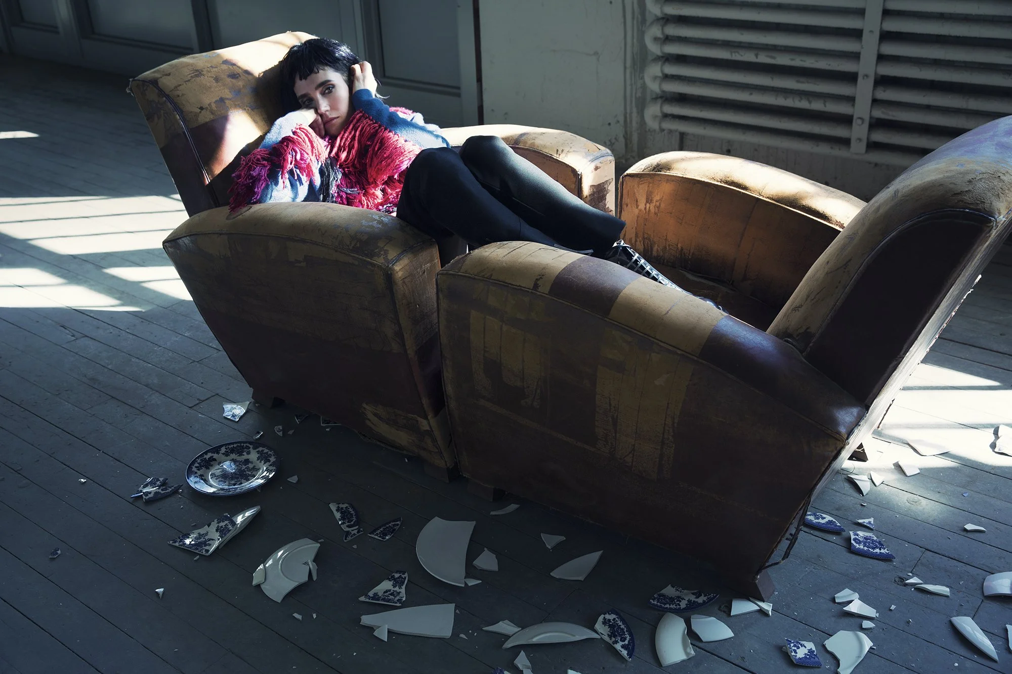 A young woman with short dark hair lying on a worn, vintage sofa in a room with sunlight streaming through the window. The floor is scattered with broken ceramic dishes and plates.