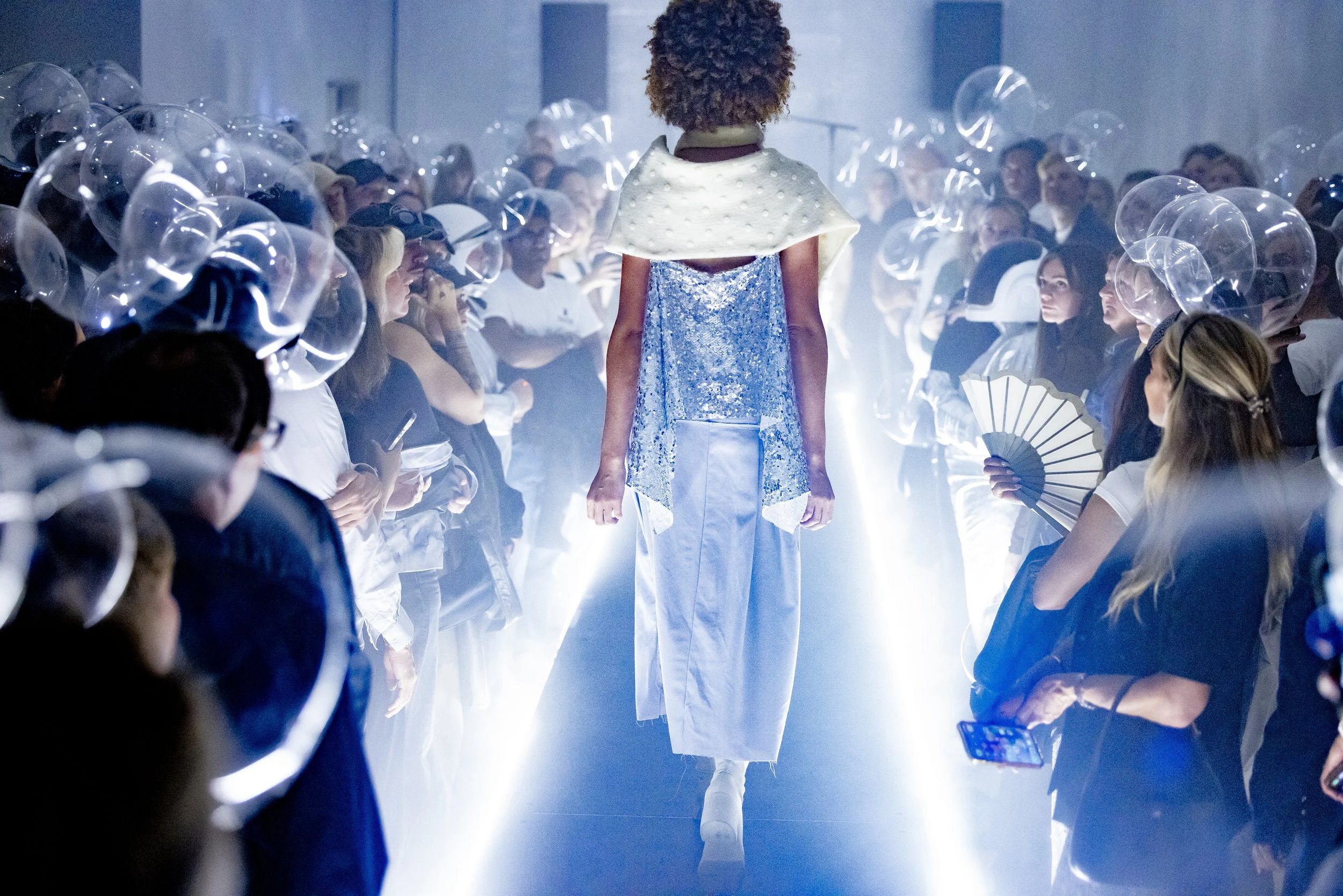 Fashion runway model walking down runway surrounded by audience members with transparent balloons, some taking photos, in a high-tech, futuristic setting with dramatic lighting.