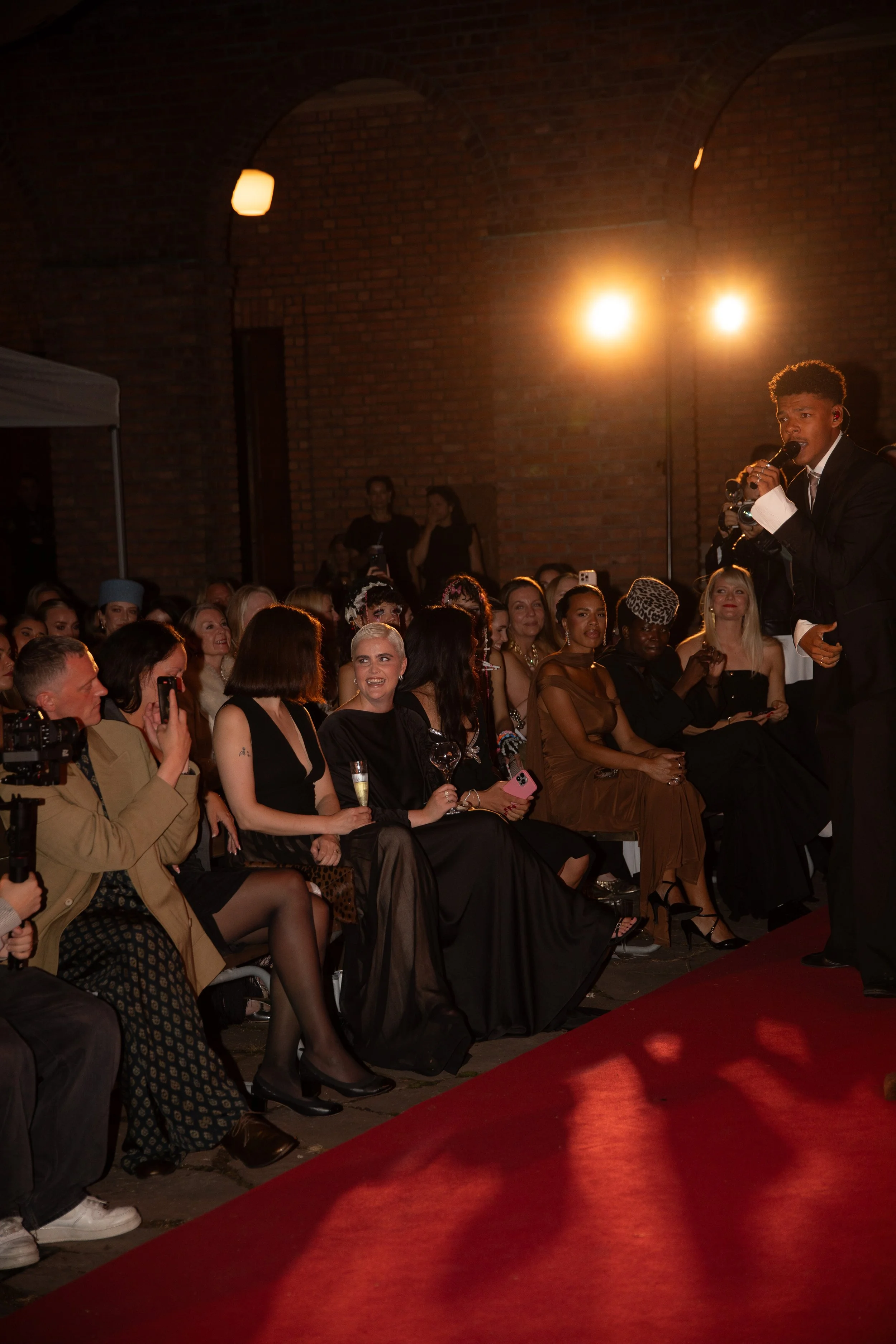 An indoor event with a seated audience watching a performer who is playing the saxophone. The event is illuminated with warm lighting and a brick wall backdrop.
