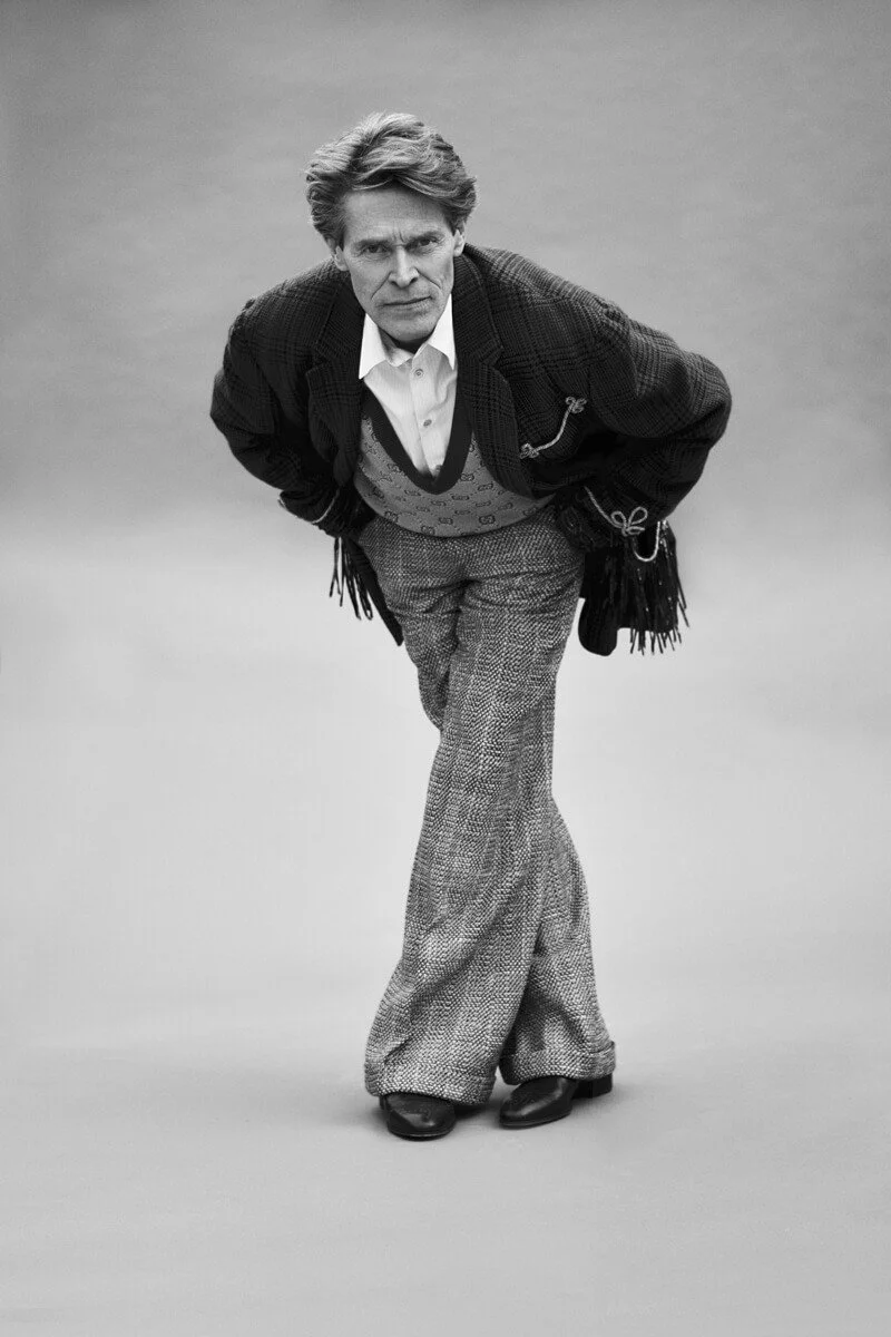 Black and white photo of a man with styled hair leaning forward with hands behind his back, wearing a checkered blazer, patterned high-waisted trousers, and a patterned sweater vest.