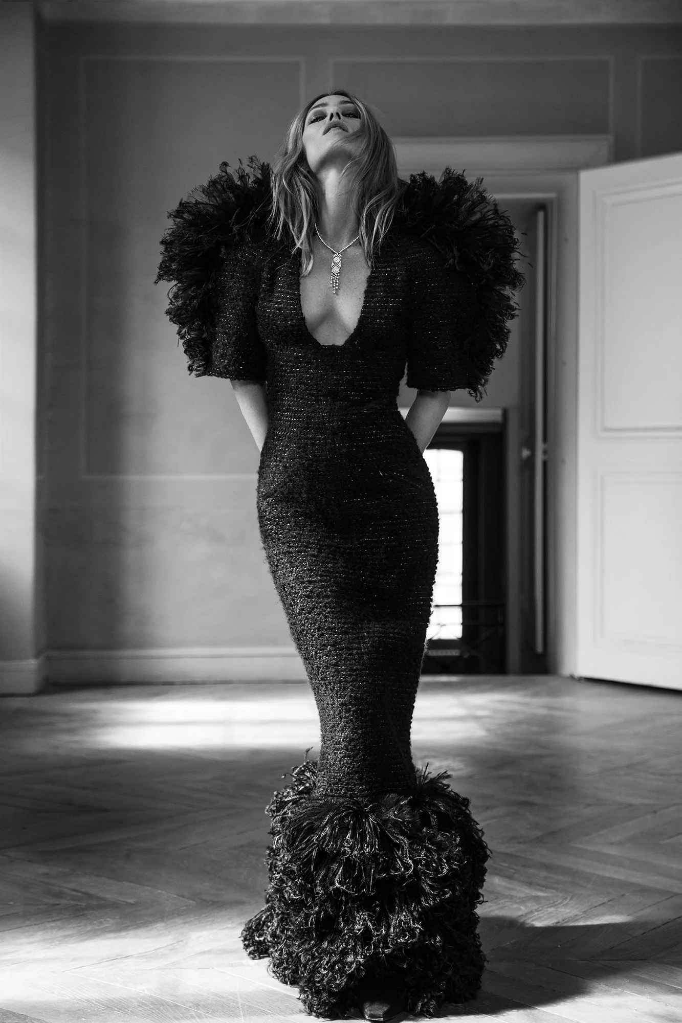 A woman wearing a black, textured, backless gown with feathered details on the shoulders and at the bottom. She is standing indoors with her head tilted back and hands behind her back.