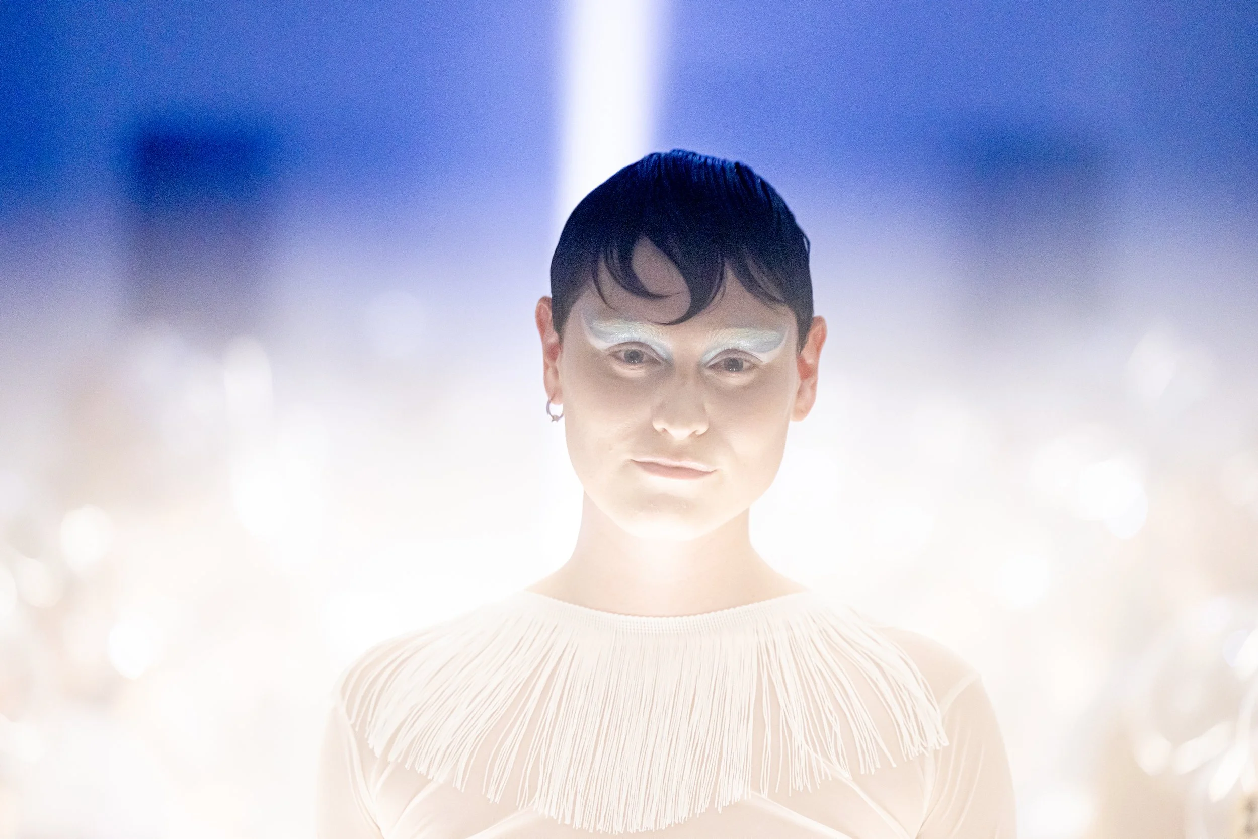 A person with a focused expression, wearing white makeup and a fringed top, with a luminous, blurred background