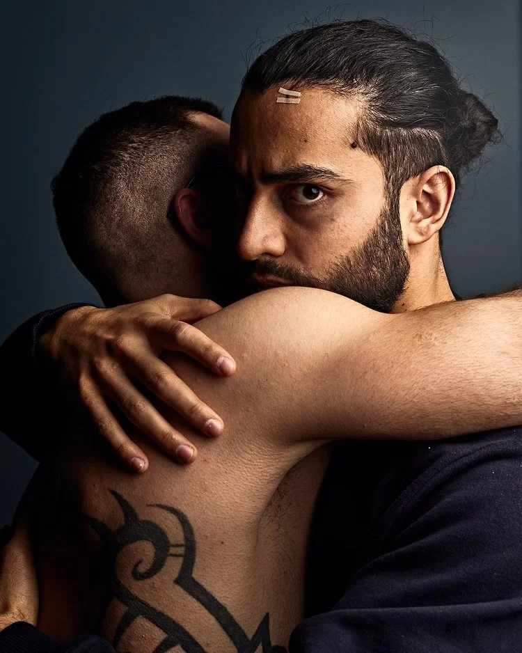 Two men hugging, one with a beard and long hair, the other with a shaved head, in an intimate embrace. The man with long hair has a serious expression and visible tattoos on his arm.