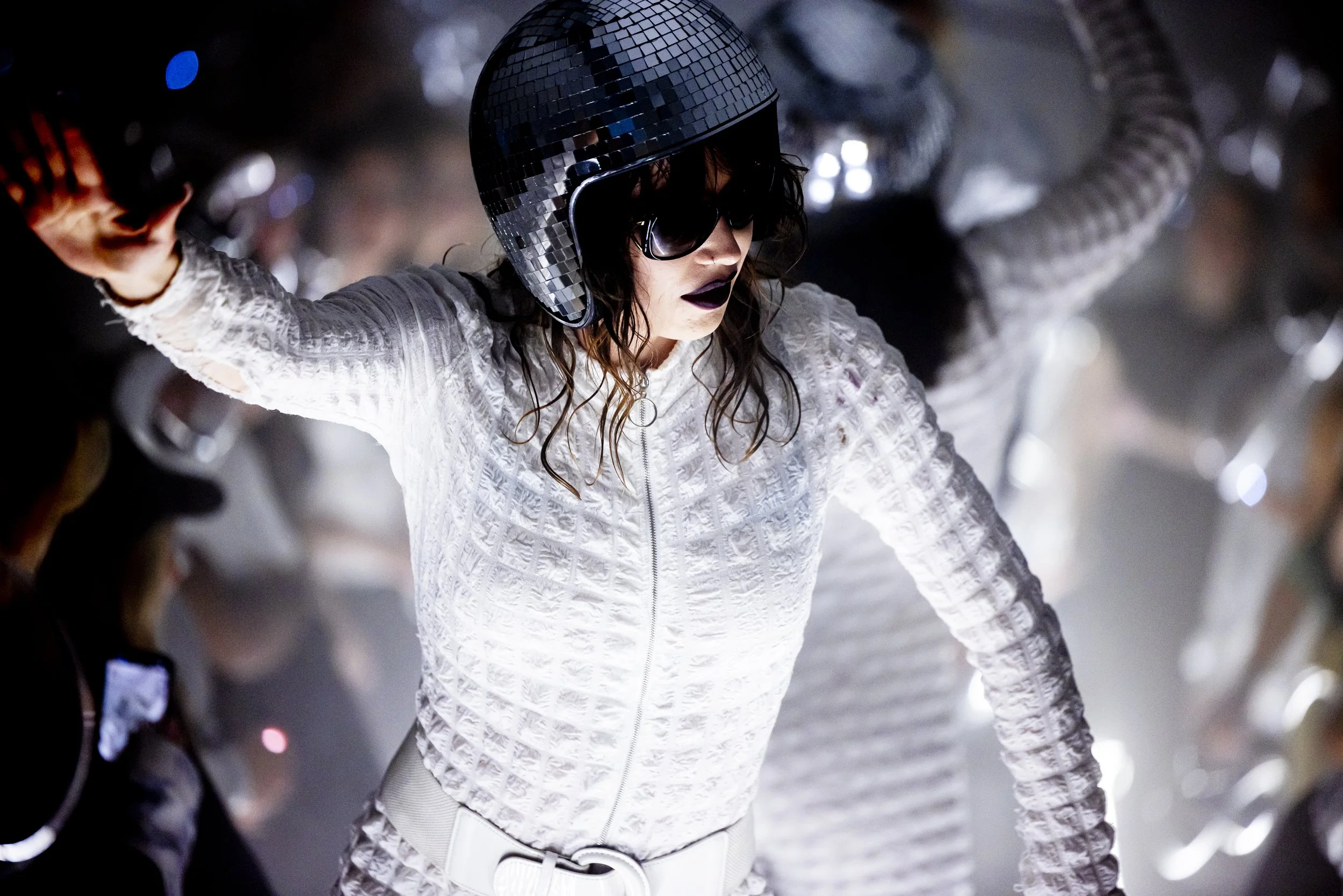 A woman wearing a quilted white jacket, black helmet with a mirror-like surface, and dark sunglasses, riding a motorcycle in a dimly lit environment.
