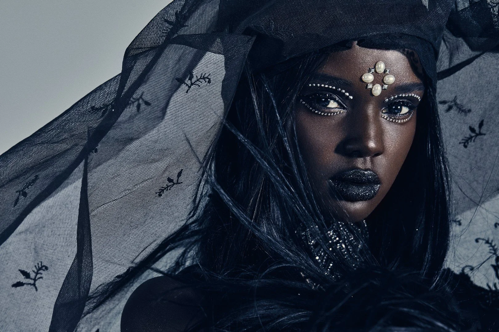 A woman with dark skin and dramatic makeup, wearing black lipstick, sequined eye makeup, and a pearl-studded forehead decoration, is partially covered by a black sheer veil with small branch and leaf patterns.