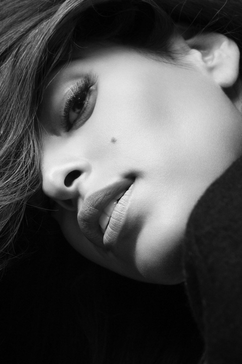 Close-up black and white photo of a woman's face, showing her eye, lips, and a small beauty mark, with her hair partially covering her face.