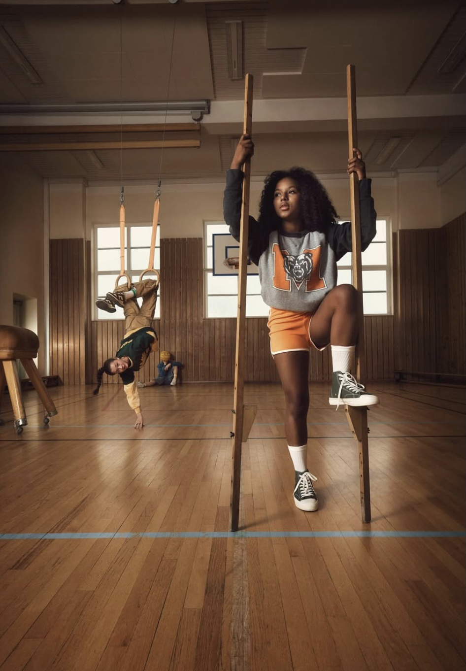 A girl dressed in sports attire practicing gymnastics on gymnastic stilts, with two girls behind her, one doing a handstand and the other sitting against a wooden wall in a gymnasium.