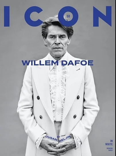 Black and white photo of a man with styled hair, wearing a white suit and lacy shirt, standing with his hands in front. The background has large blue text that reads 'ICON' and the man is labeled 'WILLEM DAFOE' in bold blue letters.
