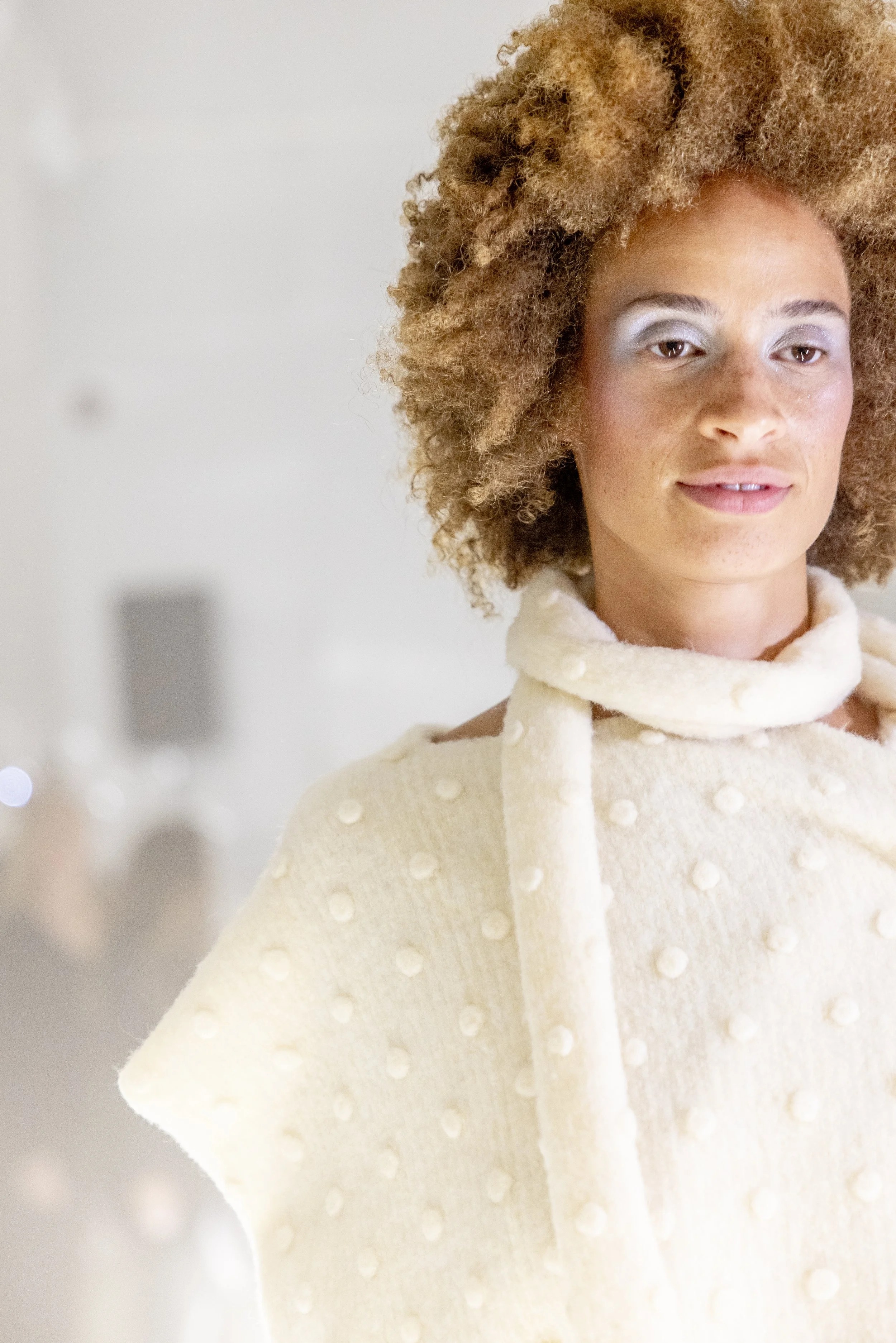 A woman with curly hair wearing a cream-colored sweater with textured dots and a scarf around her neck, looking slightly to the side.