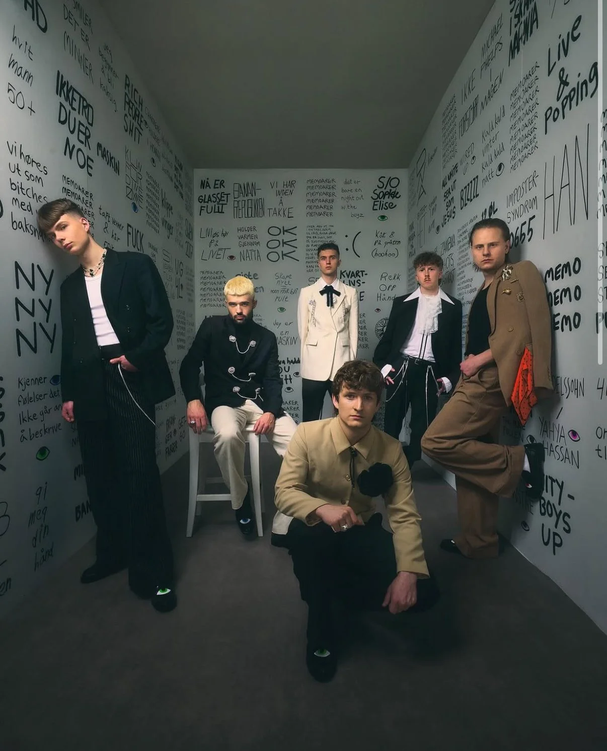 Six people in a small room with walls covered in handwritten text. Two are seated, four are standing, all wearing fashionable clothing and looking at the camera.