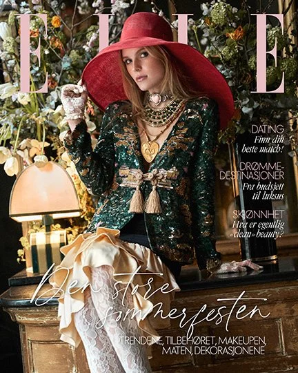 A woman with long hair wearing a large red hat, a black sequined top, layered necklaces, and a cream-colored skirt with lace and bow details. She is in a room with floral decor, a lamp, and appears on the cover of a magazine.