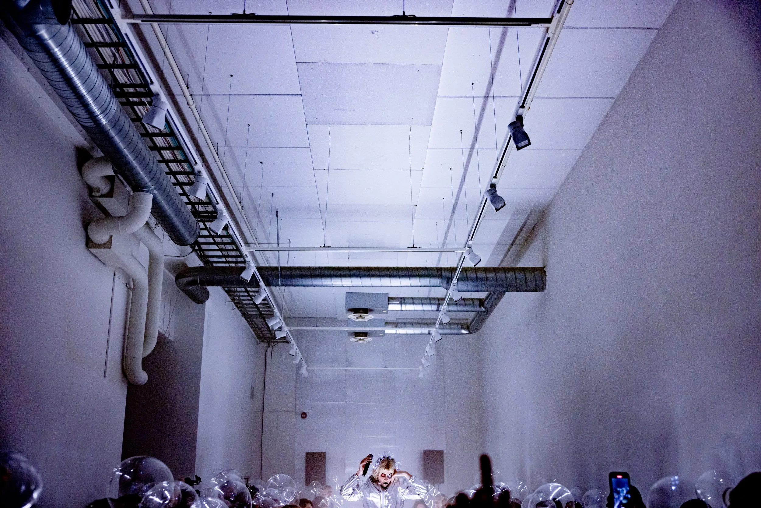A person dressed in a shiny silver outfit wearing bold makeup stands in front of a large crowd in an industrial-style room with a high ceiling, exposed ducts, and track lighting.