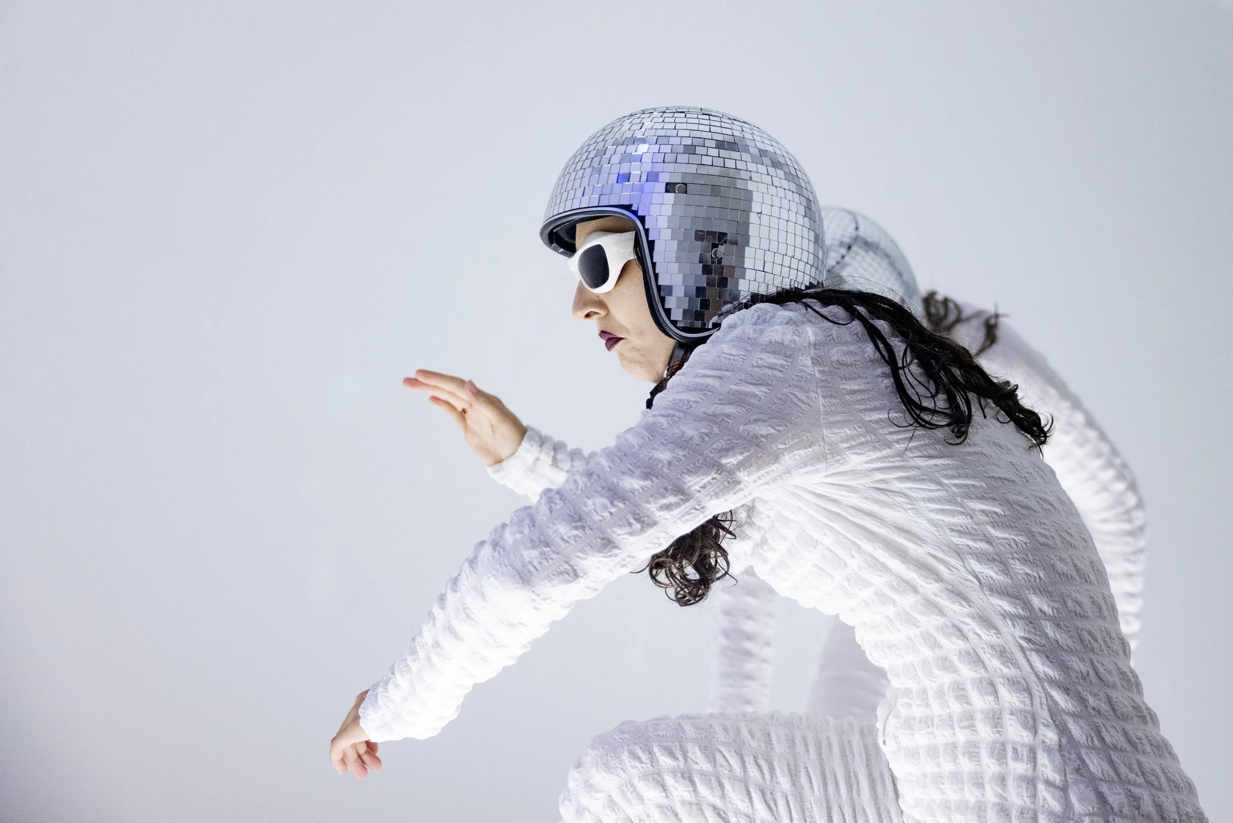 A woman wearing a white textured jumpsuit, reflective helmet covered with small square mirrors, and sunglasses, crouching and extending her arm forward in a pose against a plain light background.
