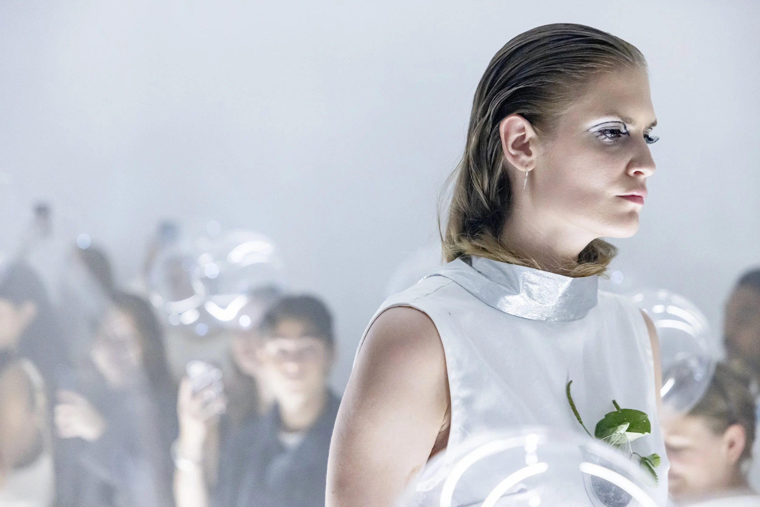 A woman with slicked-back hair and silver makeup looking to the right, wearing a futuristic white dress with metallic accents, surrounded by a blurred crowd and balloons in a bright, minimalistic setting.