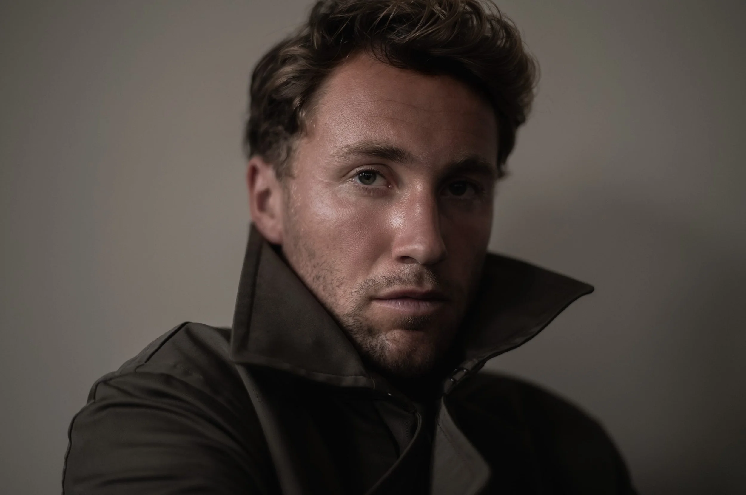 A man with short, wavy hair and a slight beard, wearing a black jacket with the collar popped, looking directly at the camera with a serious expression.