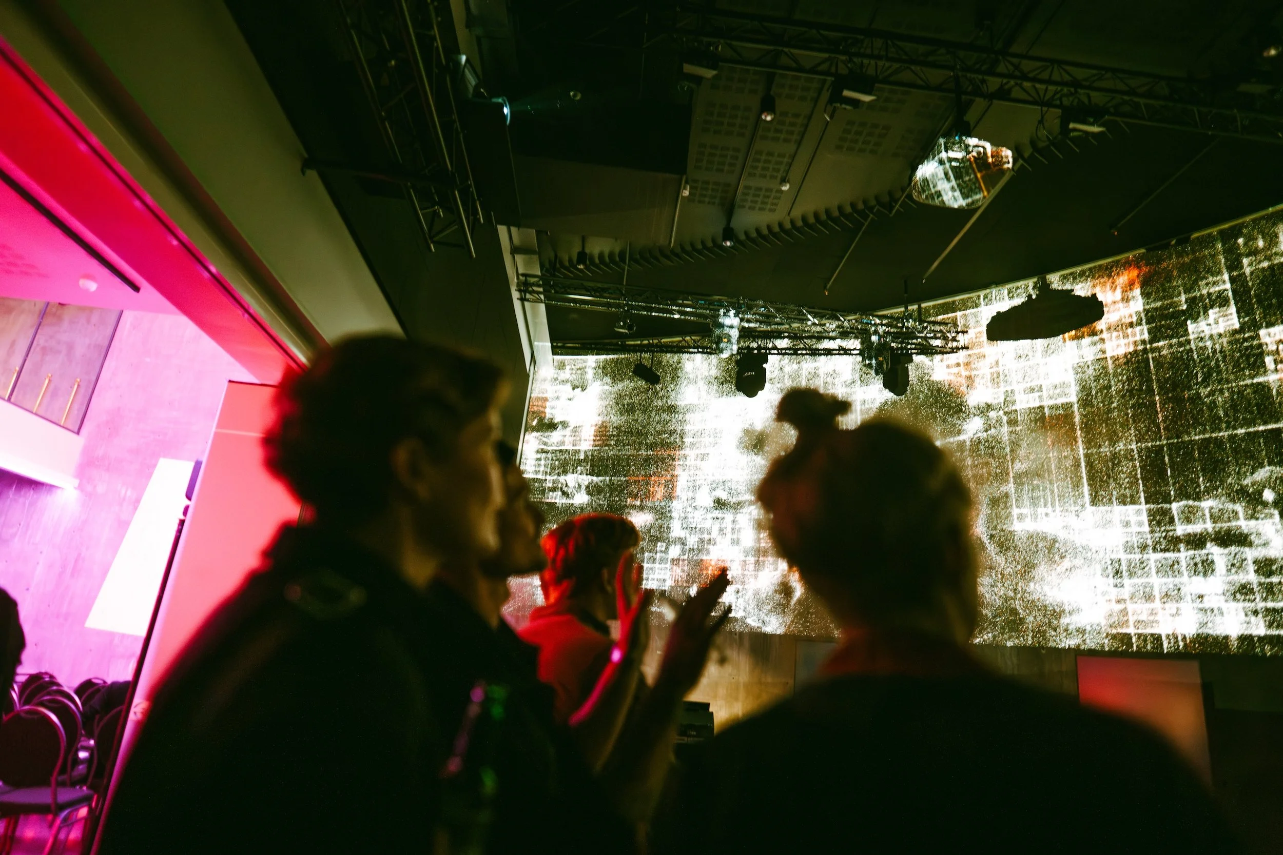 People mingling in a dimly lit venue with a large digital screen displaying abstract visuals in the background.