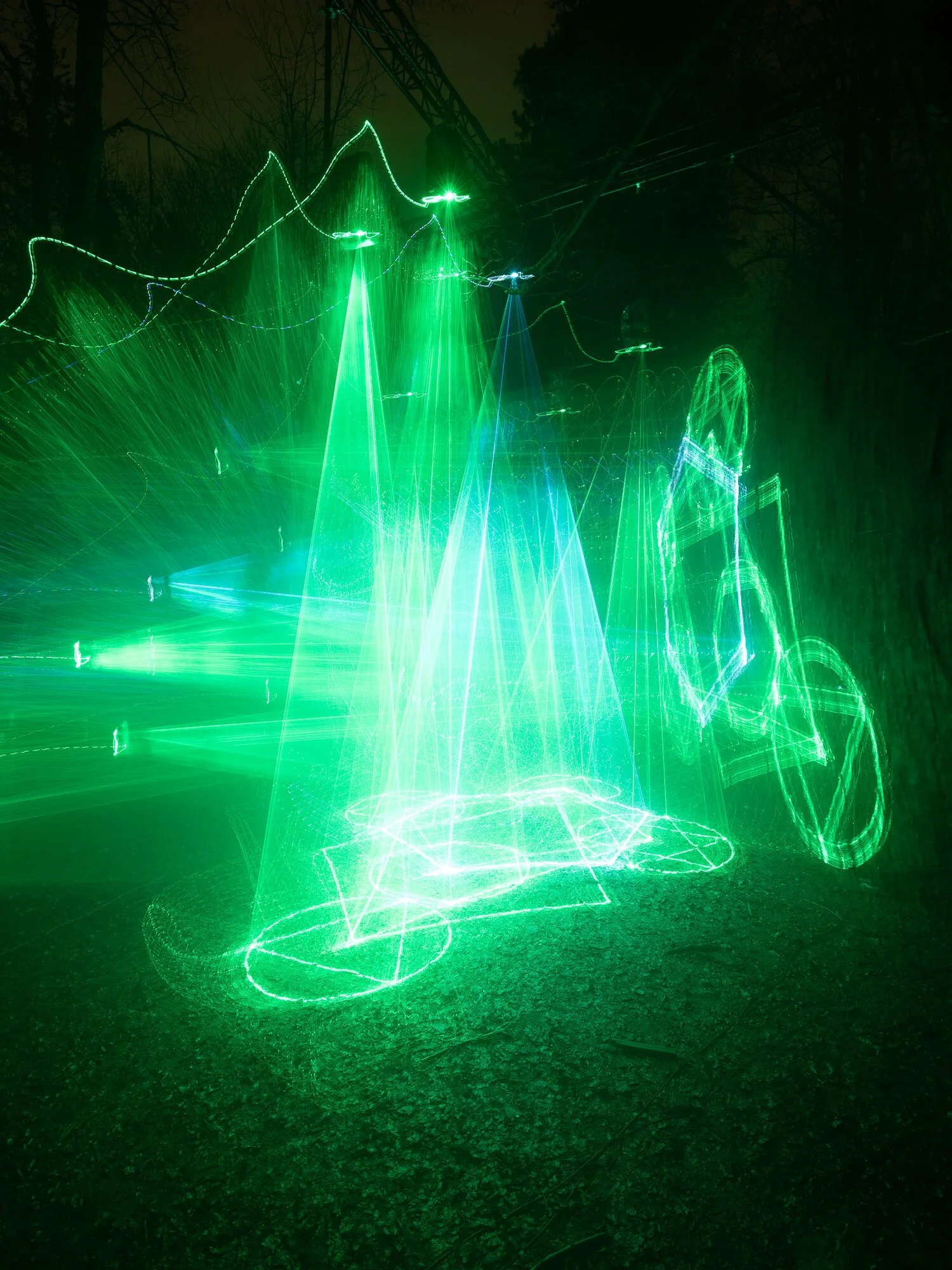 Cathedral of Ego: Light, Code, and Transcendence  MAXIME PLESCIA-BÜSCHI AND BJØRN STAAL  x COLLECTIVE OSLO Green laser light art installation with geometric shapes and lines at night, with trees and a power line structure in the background.