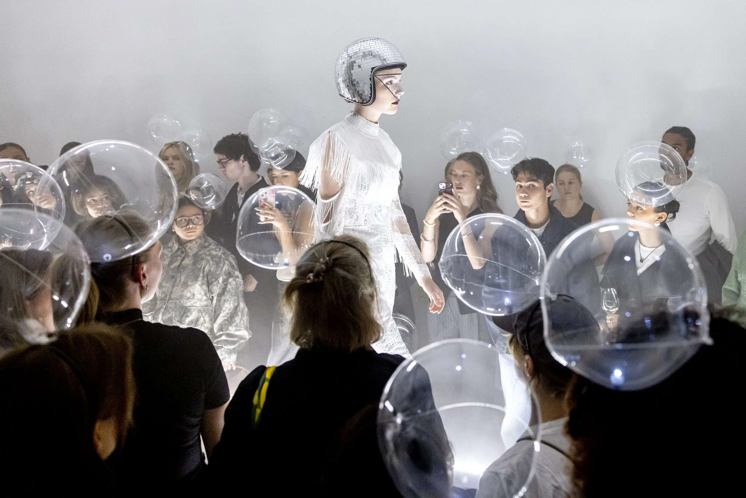 A model in a white fringed outfit and helmet walks down a runway, surrounded by an audience wearing helmets and taking photos, with a disco ball overhead.