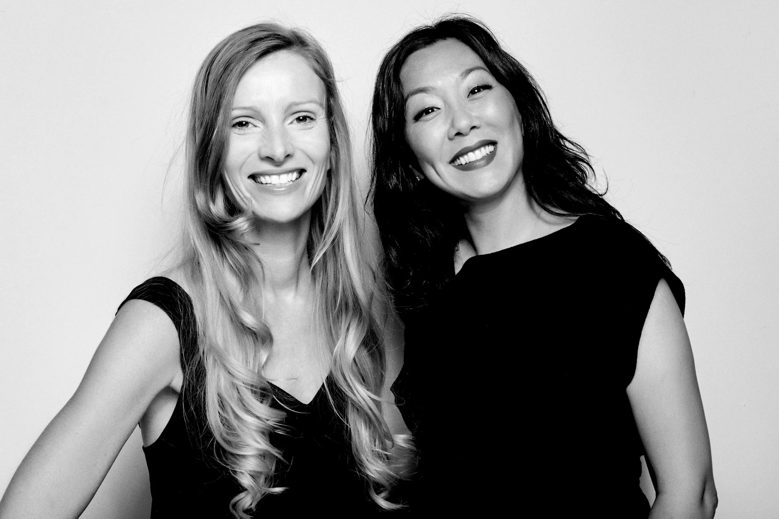 Two women smiling, one with long blonde hair and the other with long black hair, wearing black dresses, in a black-and-white photo.