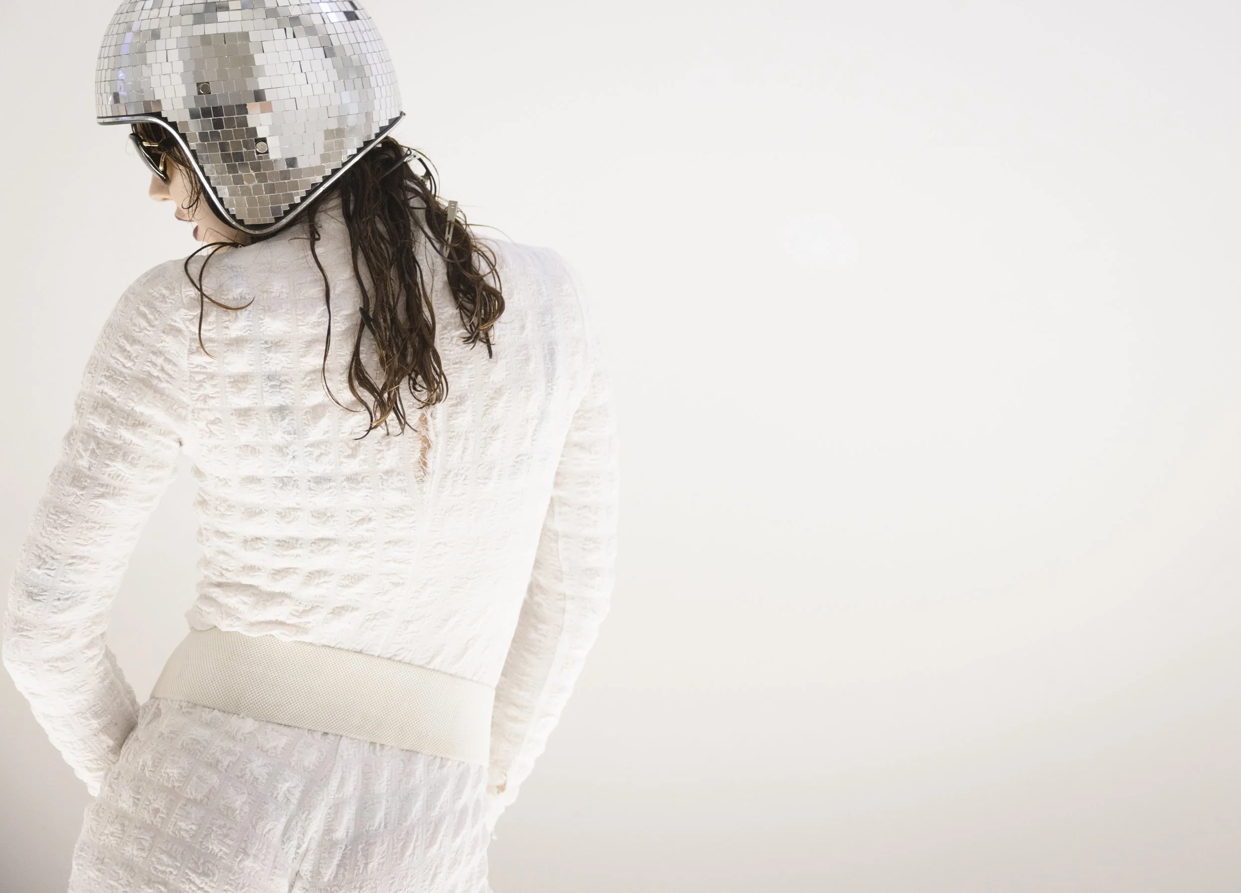 A woman with long curly hair wearing a shiny silver disco ball helmet, a white textured long-sleeve top, and matching white skirt, standing against a plain light background.