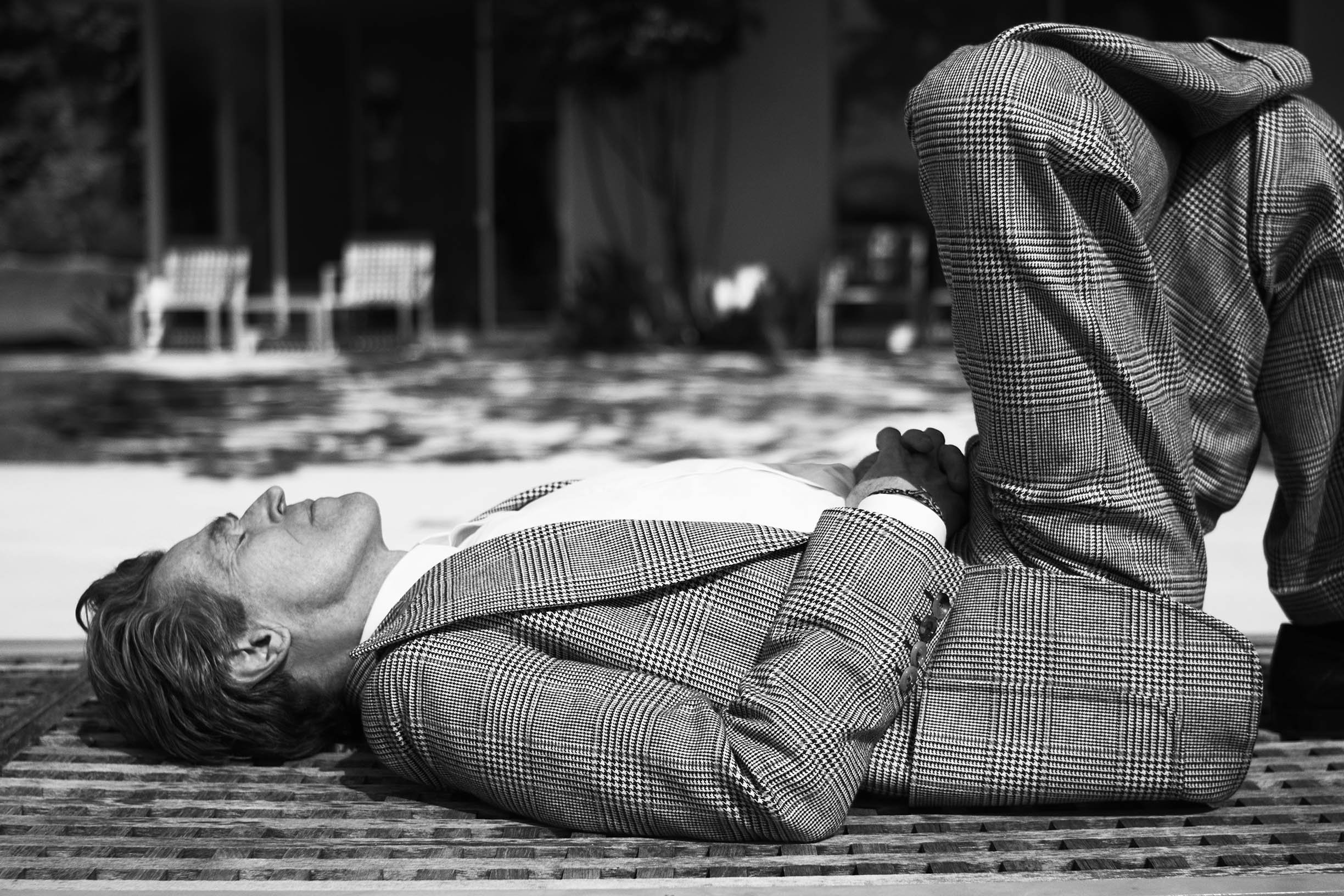 A man in a checkered suit lying on his back on a mat outdoors with a woman kneeling beside him, holding his hands, in black and white.