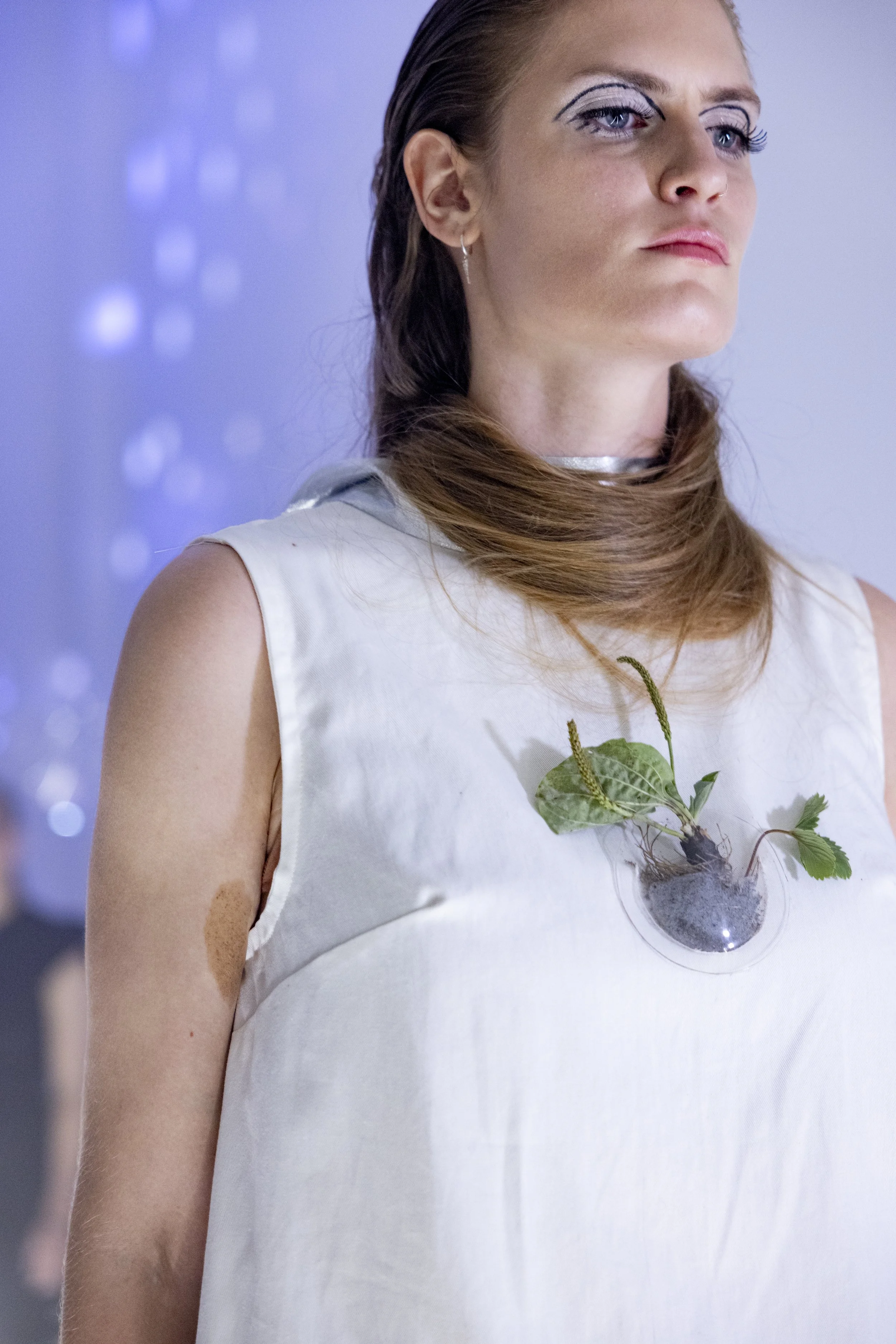 A woman with shoulder-length brown hair and makeup with bold eye lines, wearing a sleeveless white top with a clear circular pin on her chest, which contains a small plant with green leaves and soil.