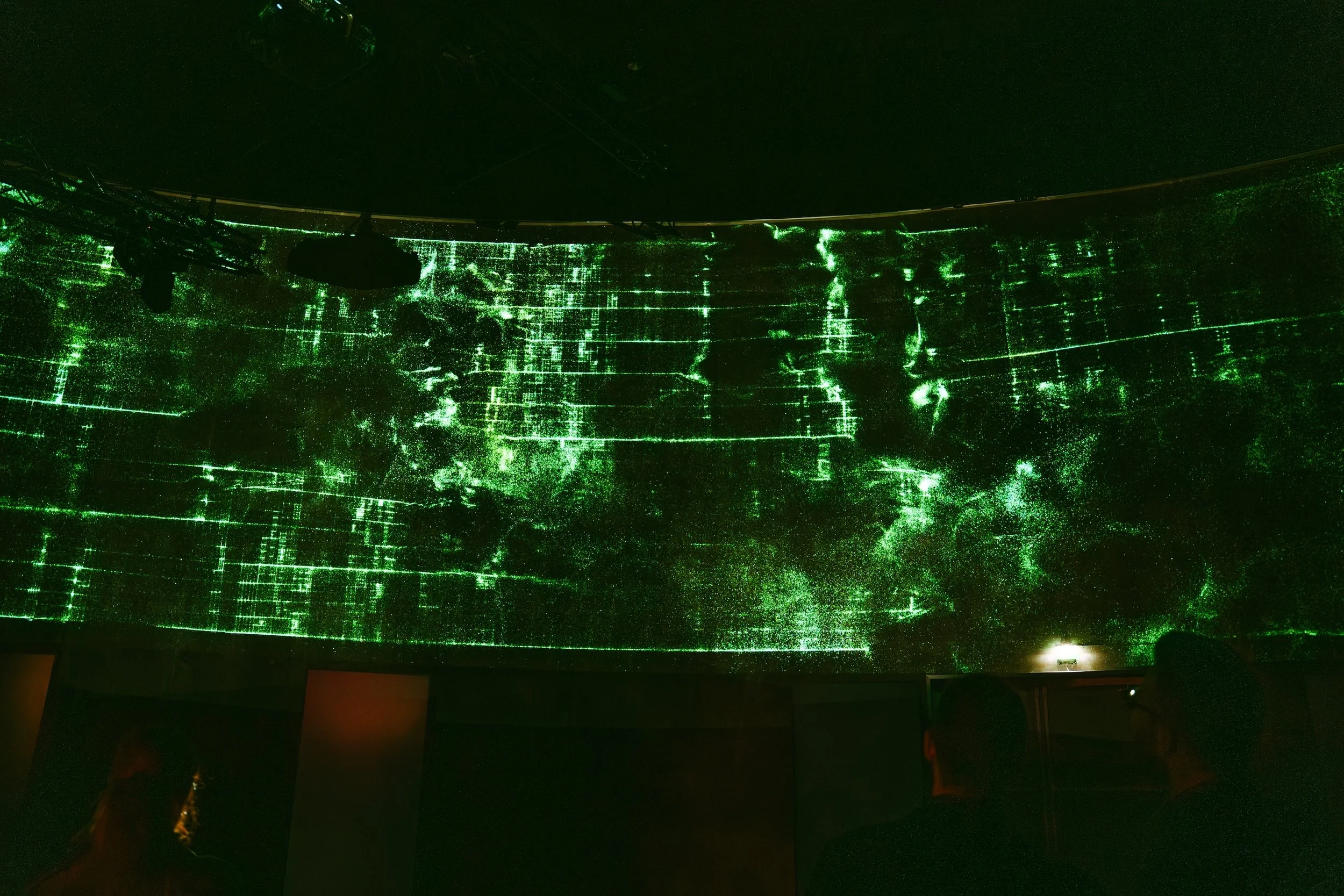 Dark room with a large screen projecting a green digital grid and map of the world, with silhouettes of people observing.