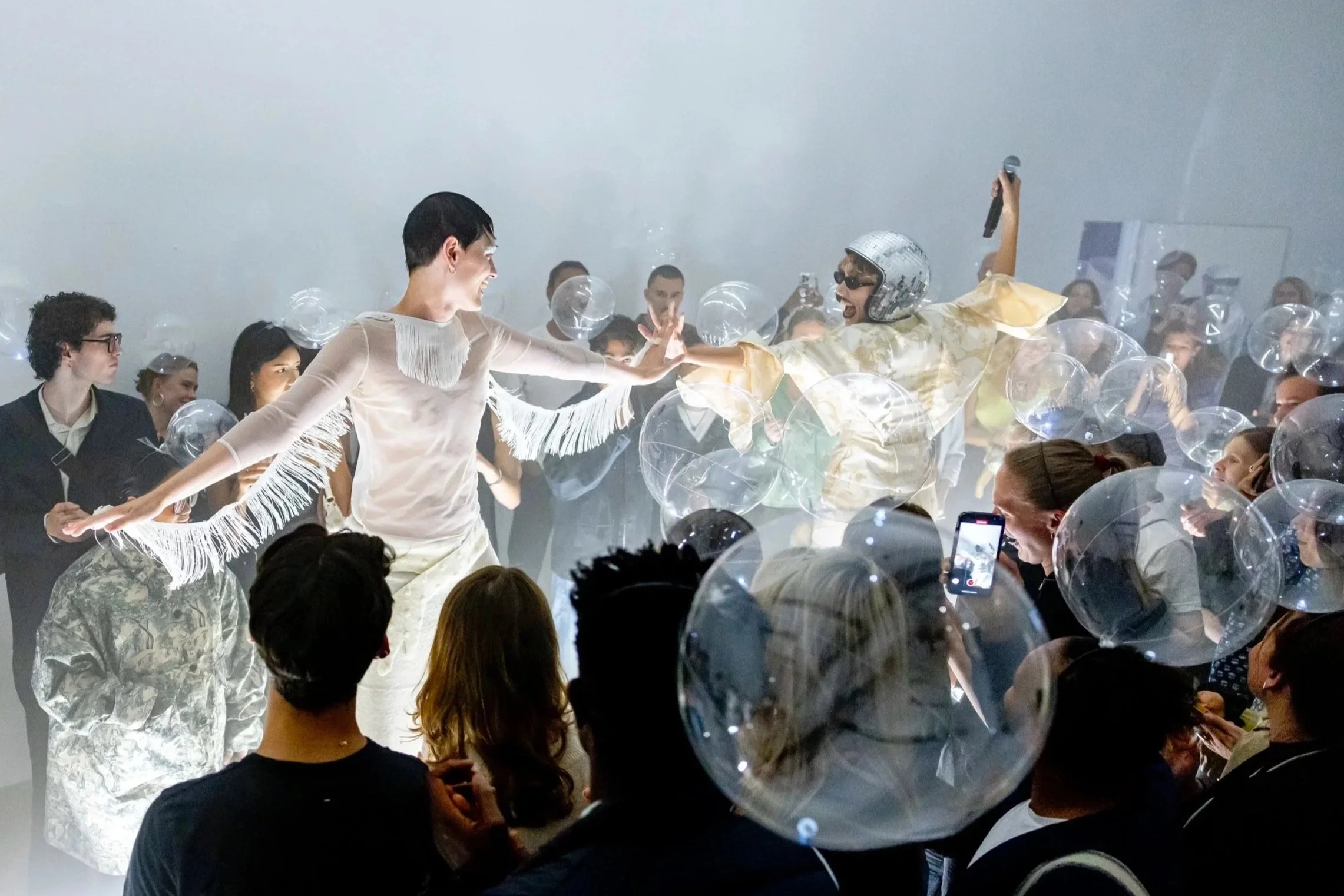 People dancing and having fun at a party surrounded by transparent balloons, with some audience members taking photos.
