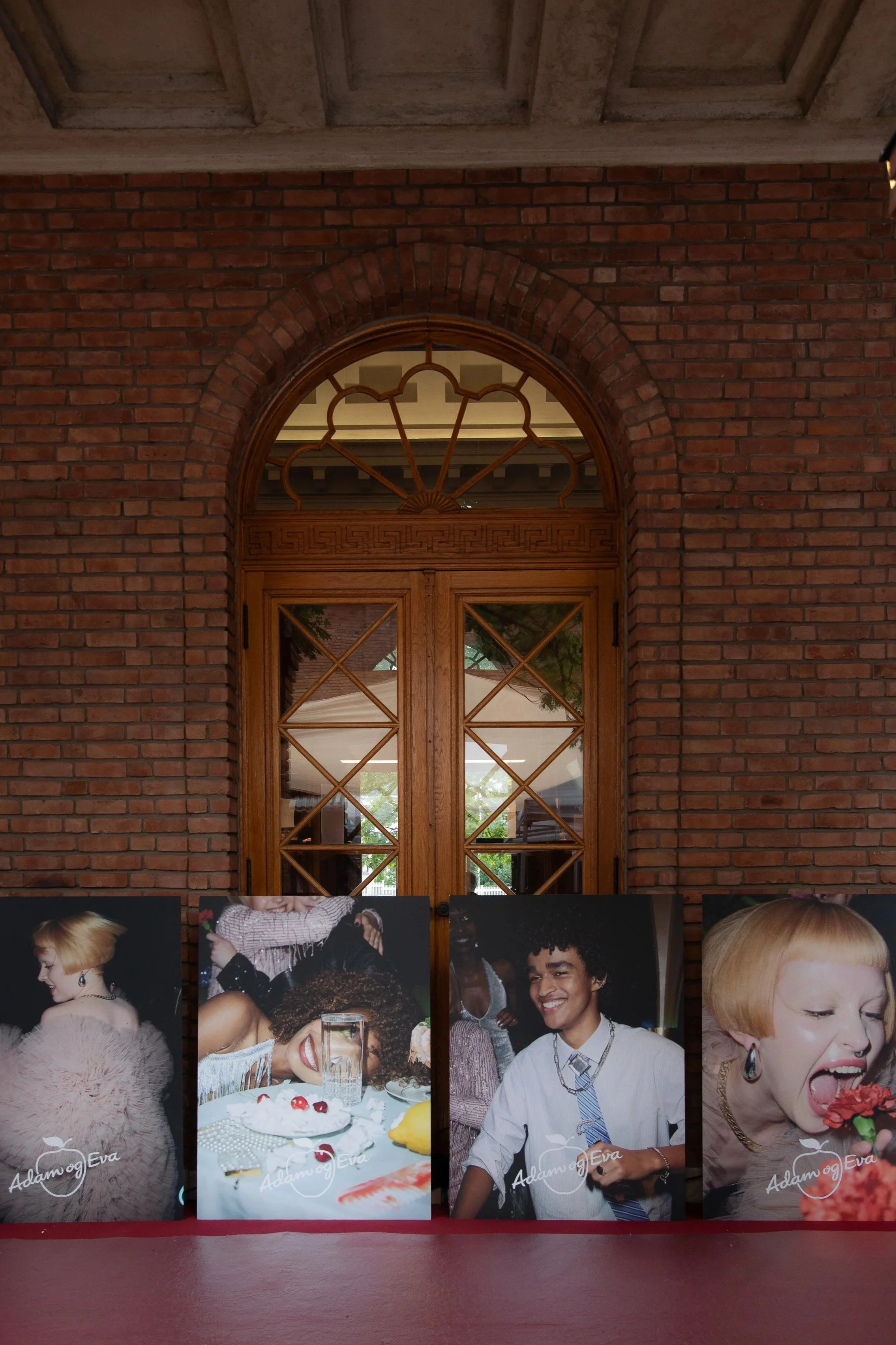 Four photographs of diverse people, including women and a man, smiling and laughing, displayed on a red surface against a brick wall with wooden double doors and an arched window above.