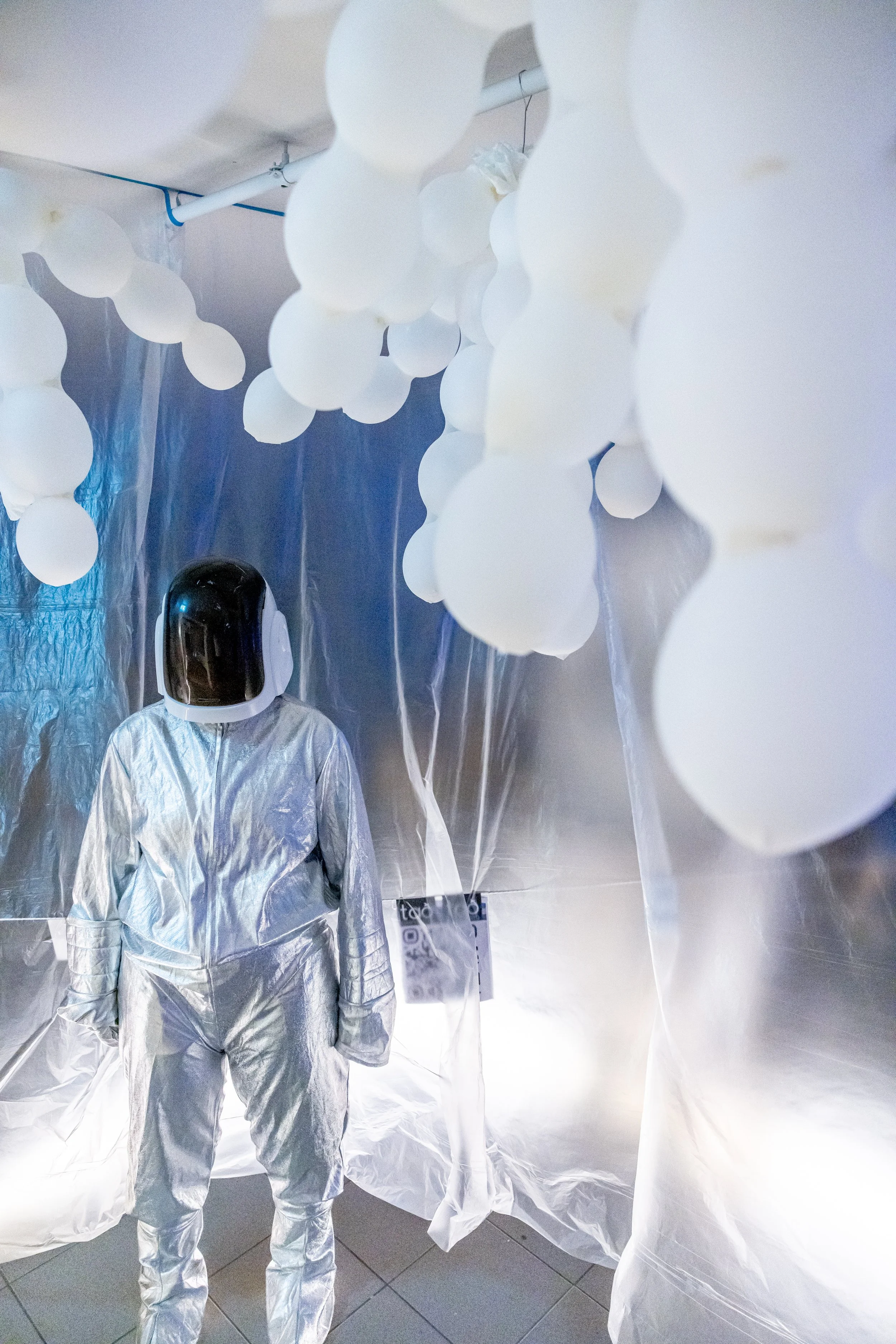 A person wearing a silver space suit and a black helmet standing in a room decorated with white balloons and silver foil walls.
