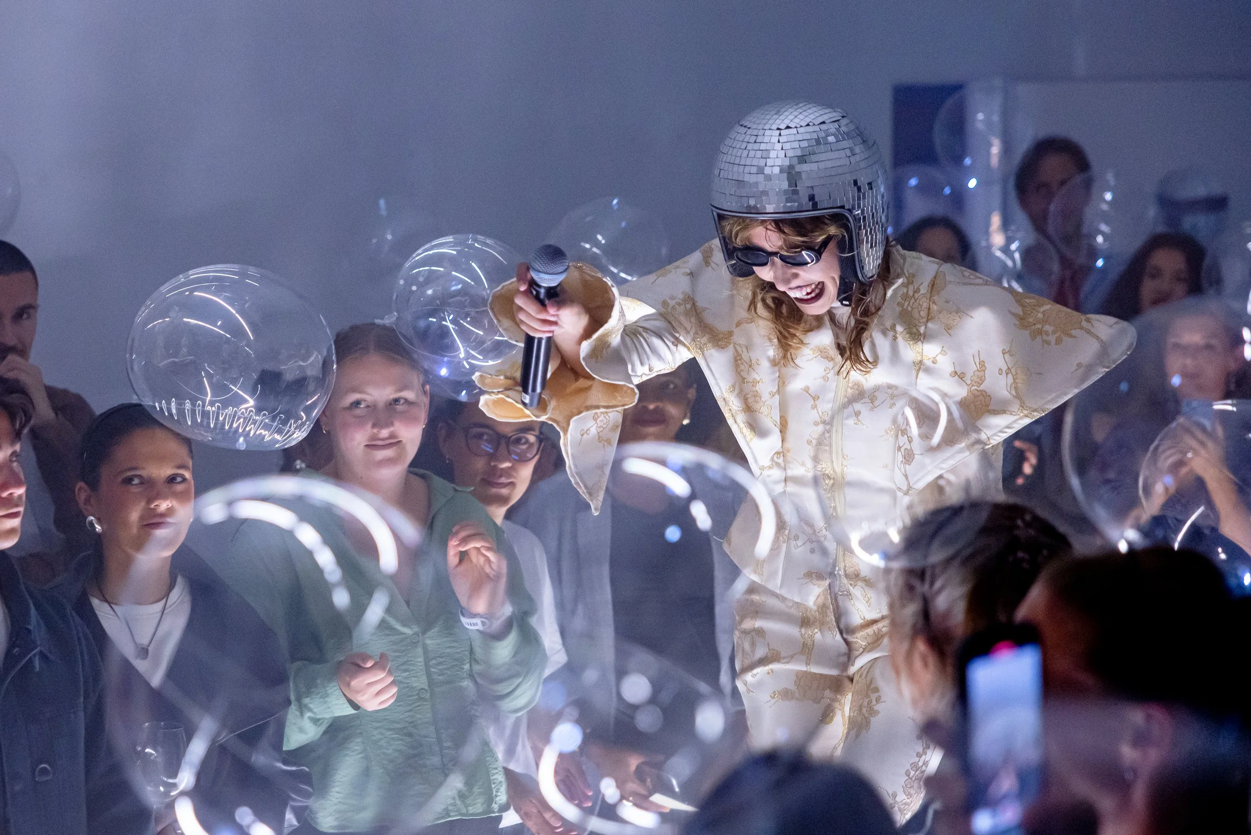 A person dressed in a shiny, gold, patterned outfit with a disco helmet, sunglasses, and holding a microphone, performing on stage with a group of audience members watching and taking photos.