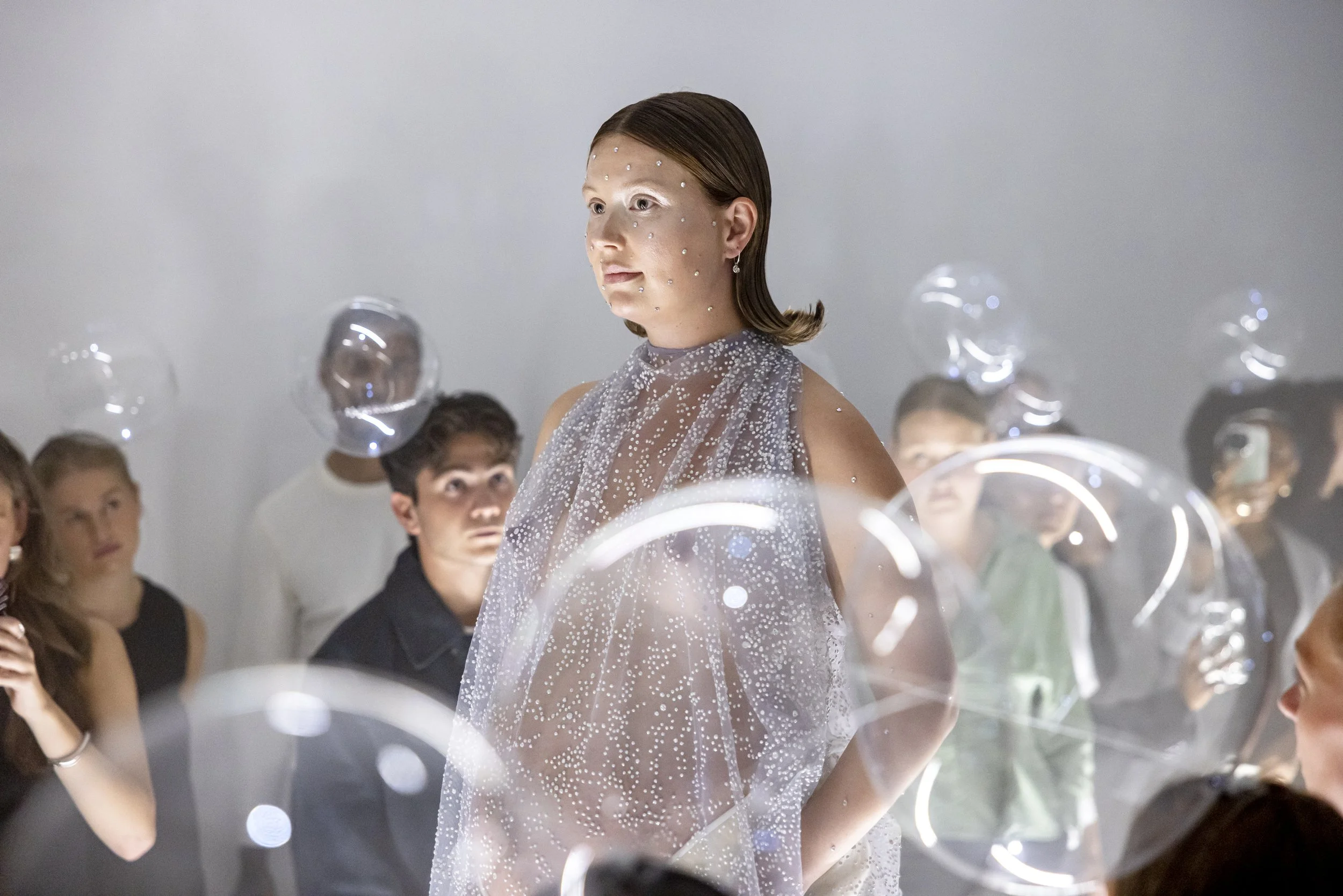 A woman wearing a sparkly, dotted dress walking through an art installation with clear balloons and a small audience watching.
