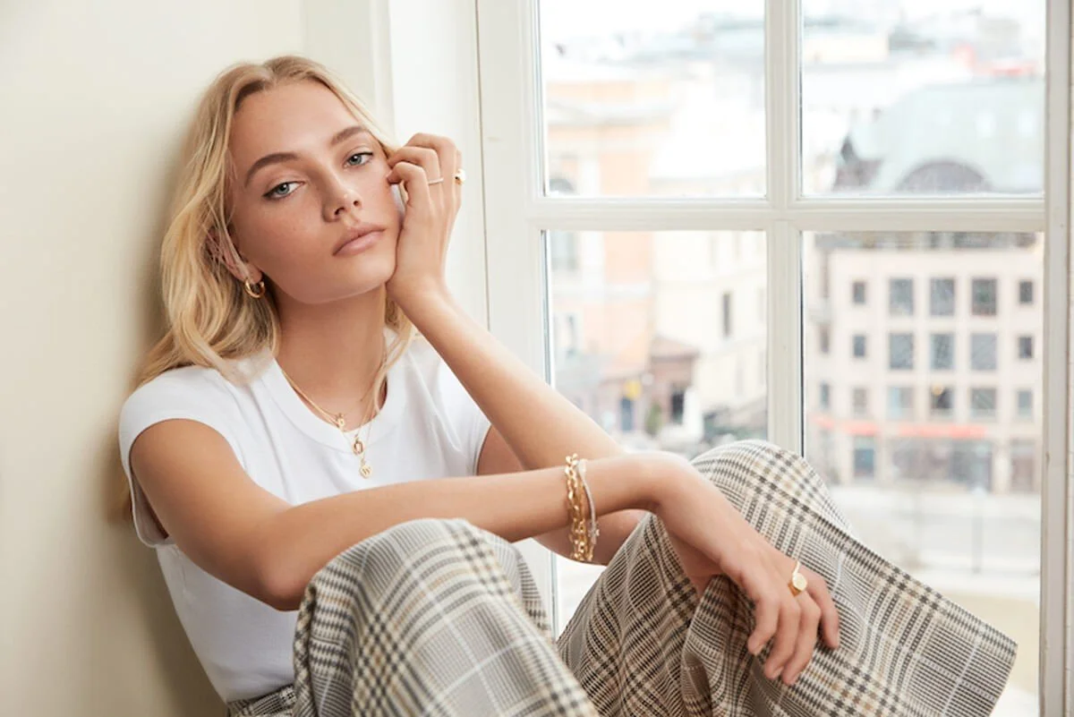 A young woman with blonde hair, dressed in a white t-shirt and plaid pants, sitting by a large window with a city view, resting her head on her hand and gazing at the camera.