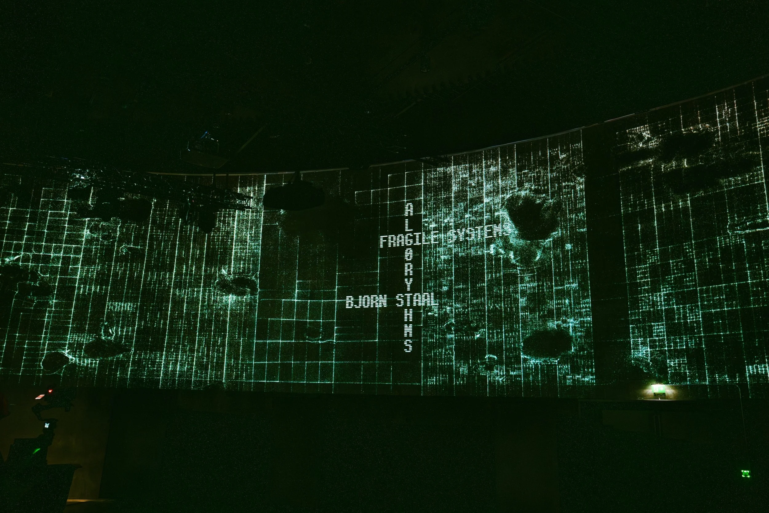 Digital projection on a large wall displaying grid lines, holes, and text including 'FRAGILE SYSTEM,' 'GBORN STEAL,' and 'H.' dark room with some lights and equipment.