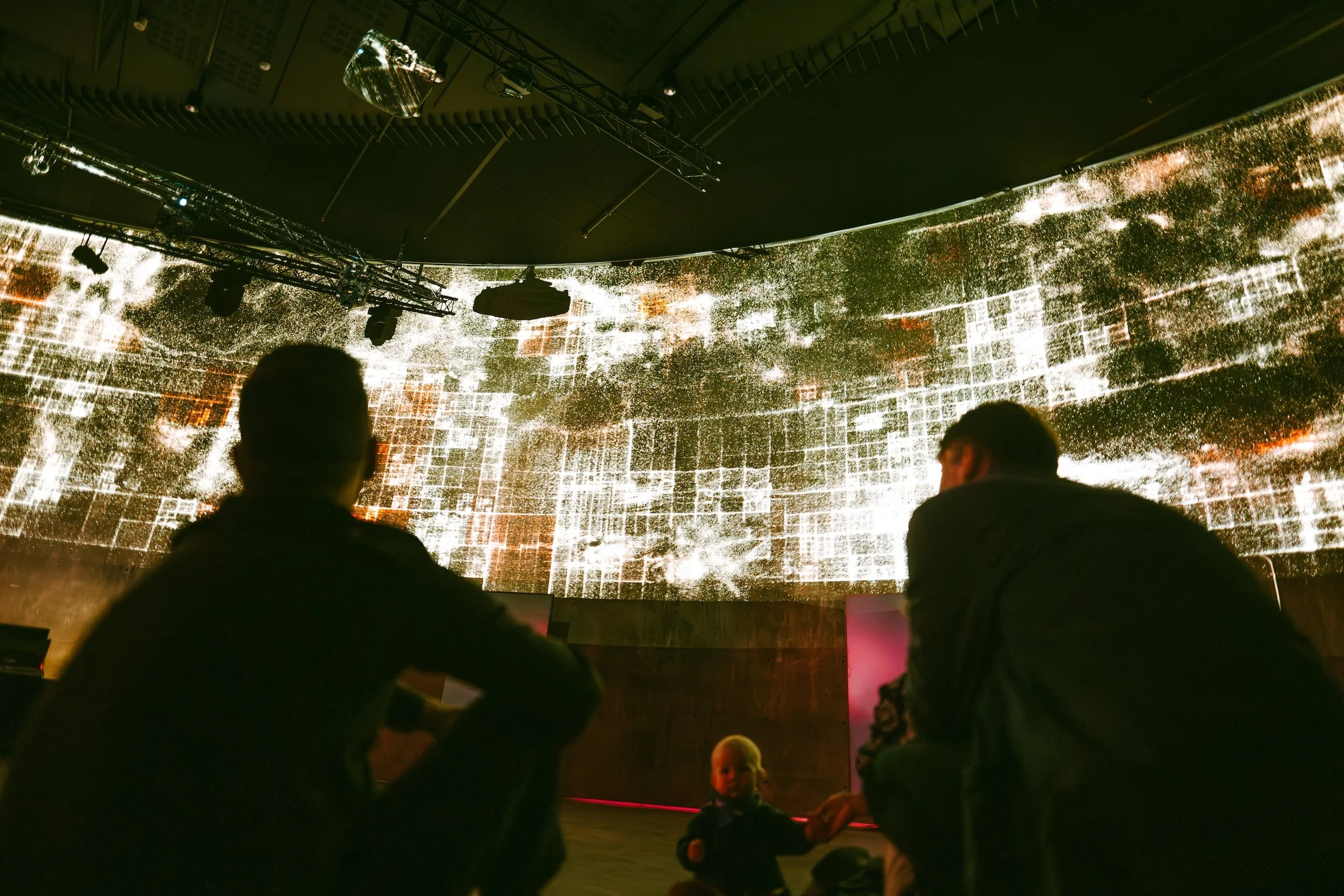 People sitting and observing a large digital art installation or display on a curved screen in a dark room, with a small child standing in front of them.