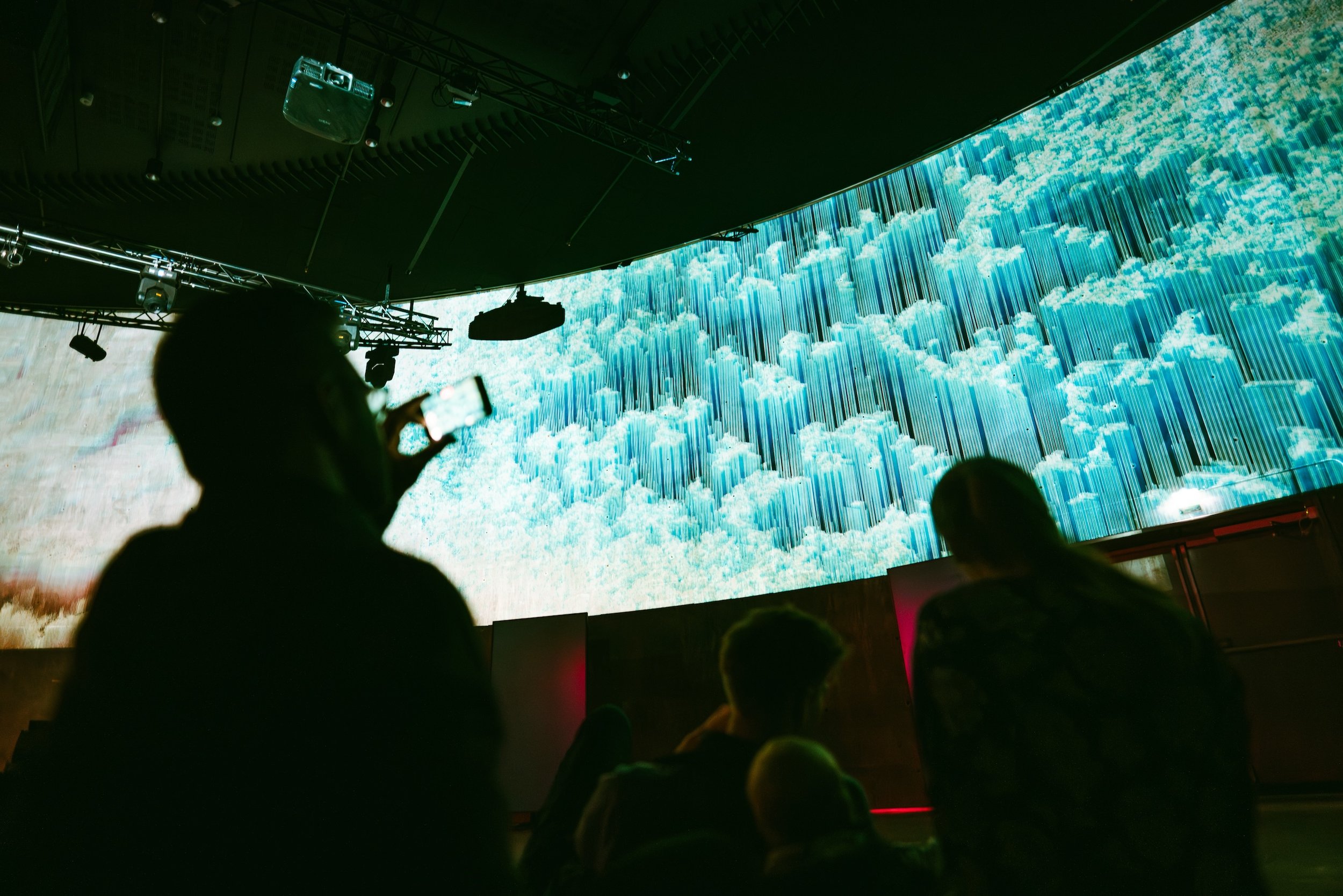 Silhouettes of people watching a large digital screen displaying blue clouds and vertical lines in a dark auditorium.