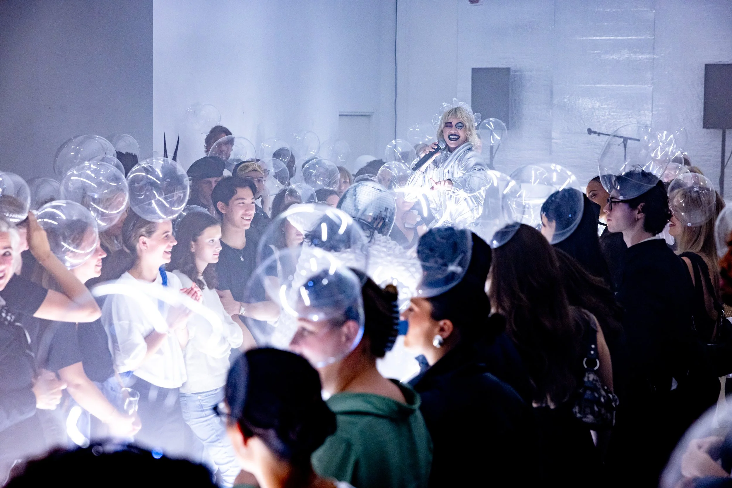 A performer with blonde hair, dark makeup, and a silver outfit singing on stage at a crowded indoor event. Audience members wear transparent helmets and are smiling and watching attentively.
