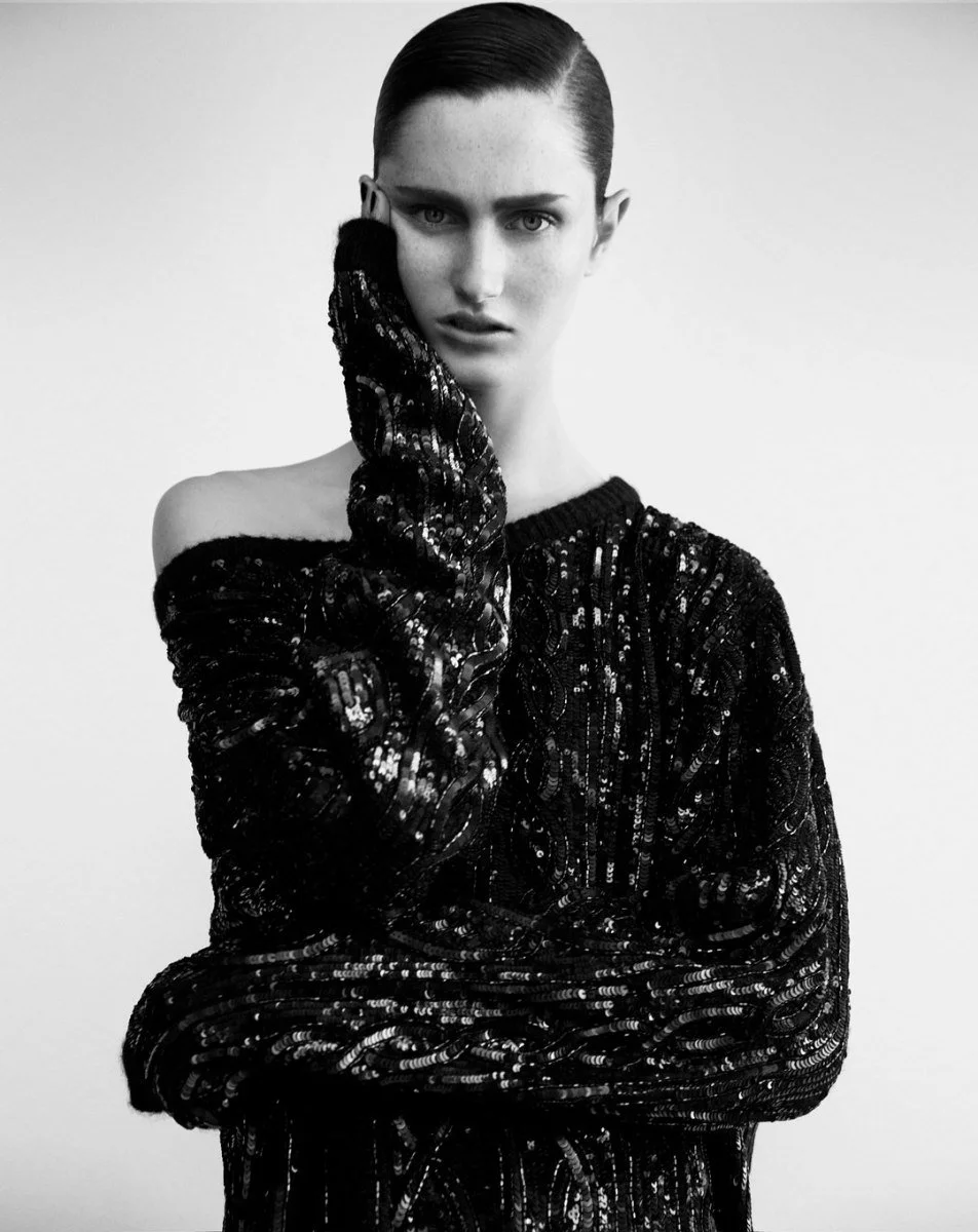 Black and white photo of a woman with slicked-back hair, wearing a dark, off-the-shoulder, sequined top, posing with her right hand on her face, looking directly at the camera.