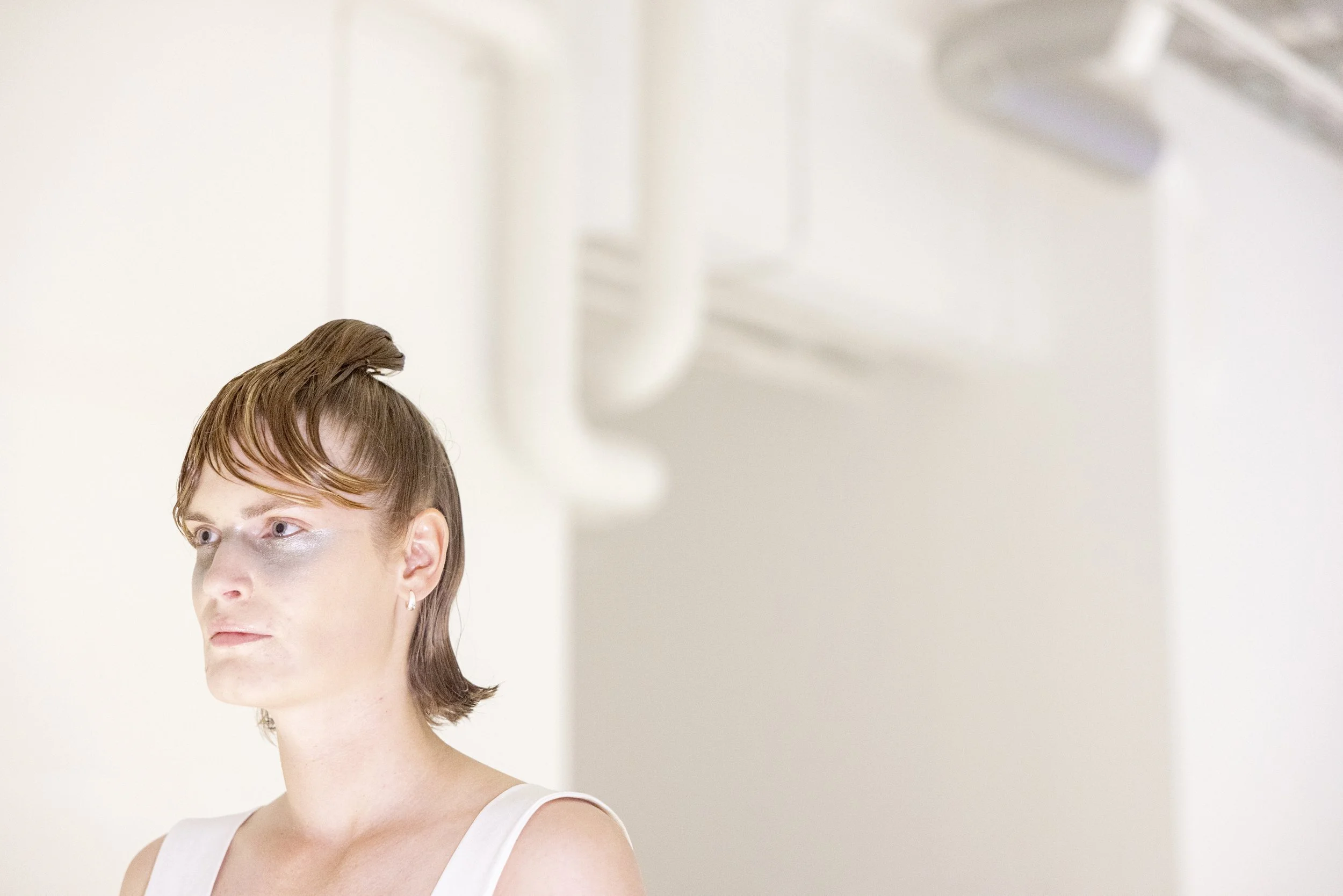 A woman with short brown hair and a white tank top looking to the side, with a metallic eyeshadow on her eyelids, standing in a brightly lit, minimalistic room.