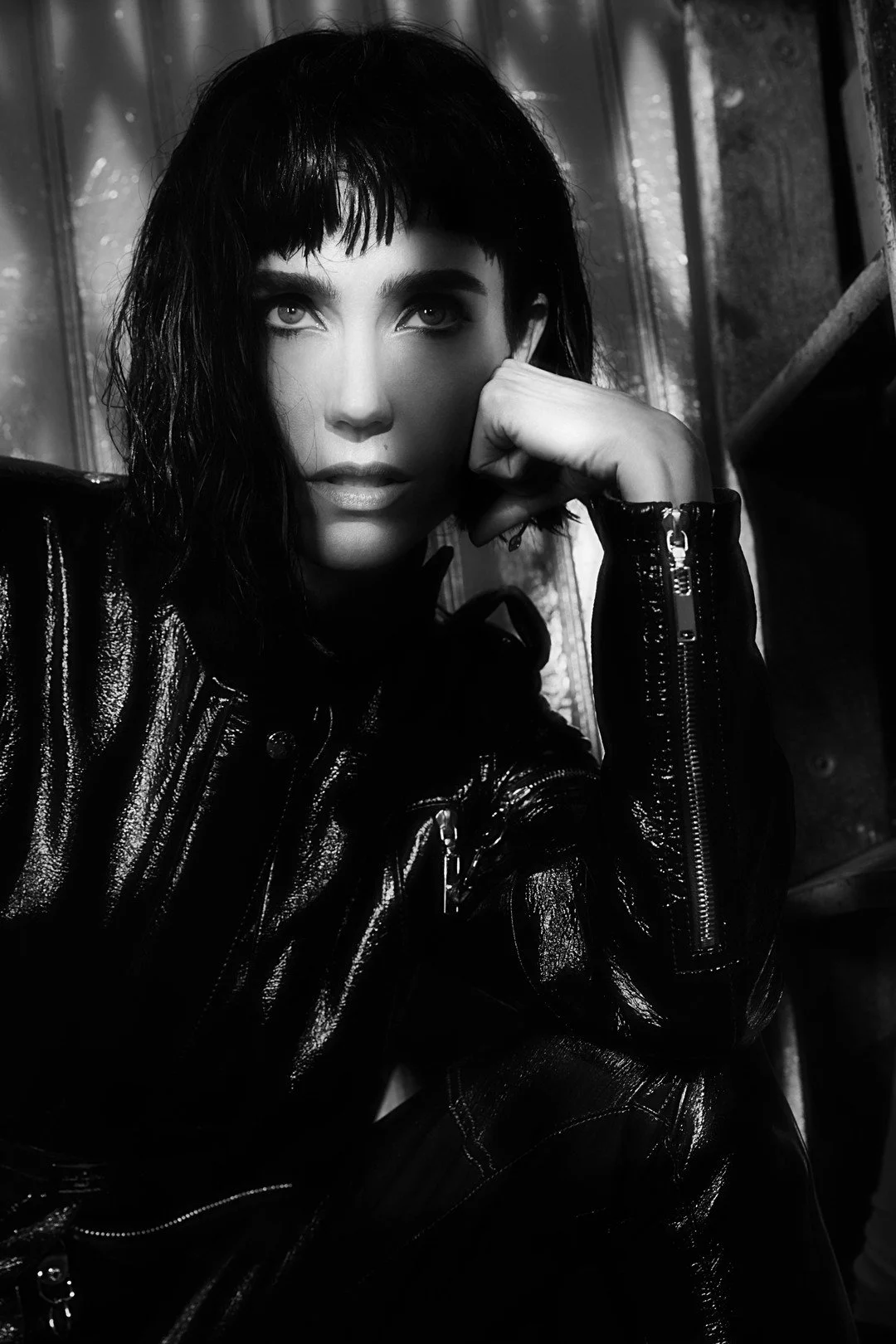 Black and white close-up photo of a woman with dark hair and bangs, wearing a shiny leather jacket, resting her head on her hand, looking at the camera.
