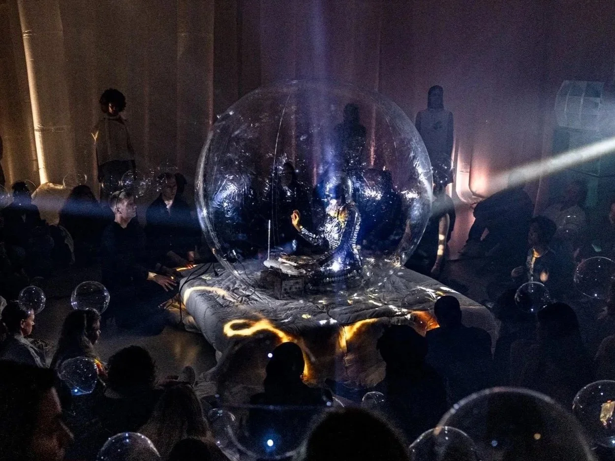 A person inside a transparent globe, possibly performing or speaking, with a dimly lit audience around them. The scene has a futuristic or artistic vibe with reflections and dramatic lighting.