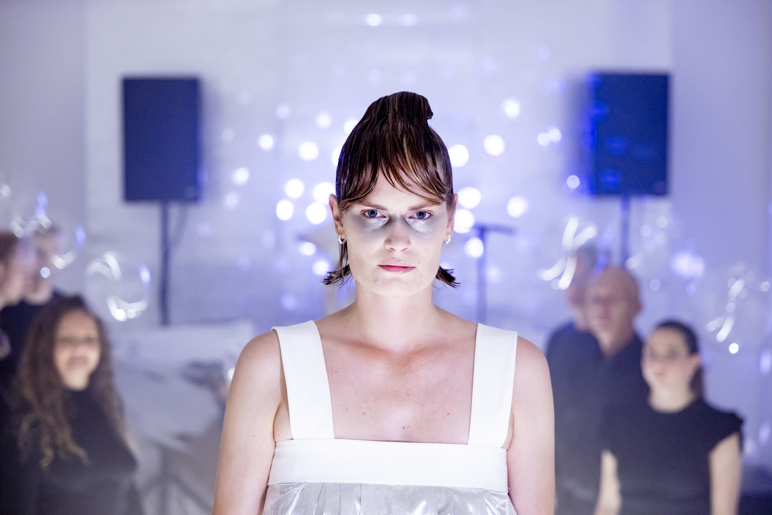 A woman with short hair, wearing a white dress, staring with a serious expression. Behind her, several people are in a blurred background at a party with festive lighting and decorations.