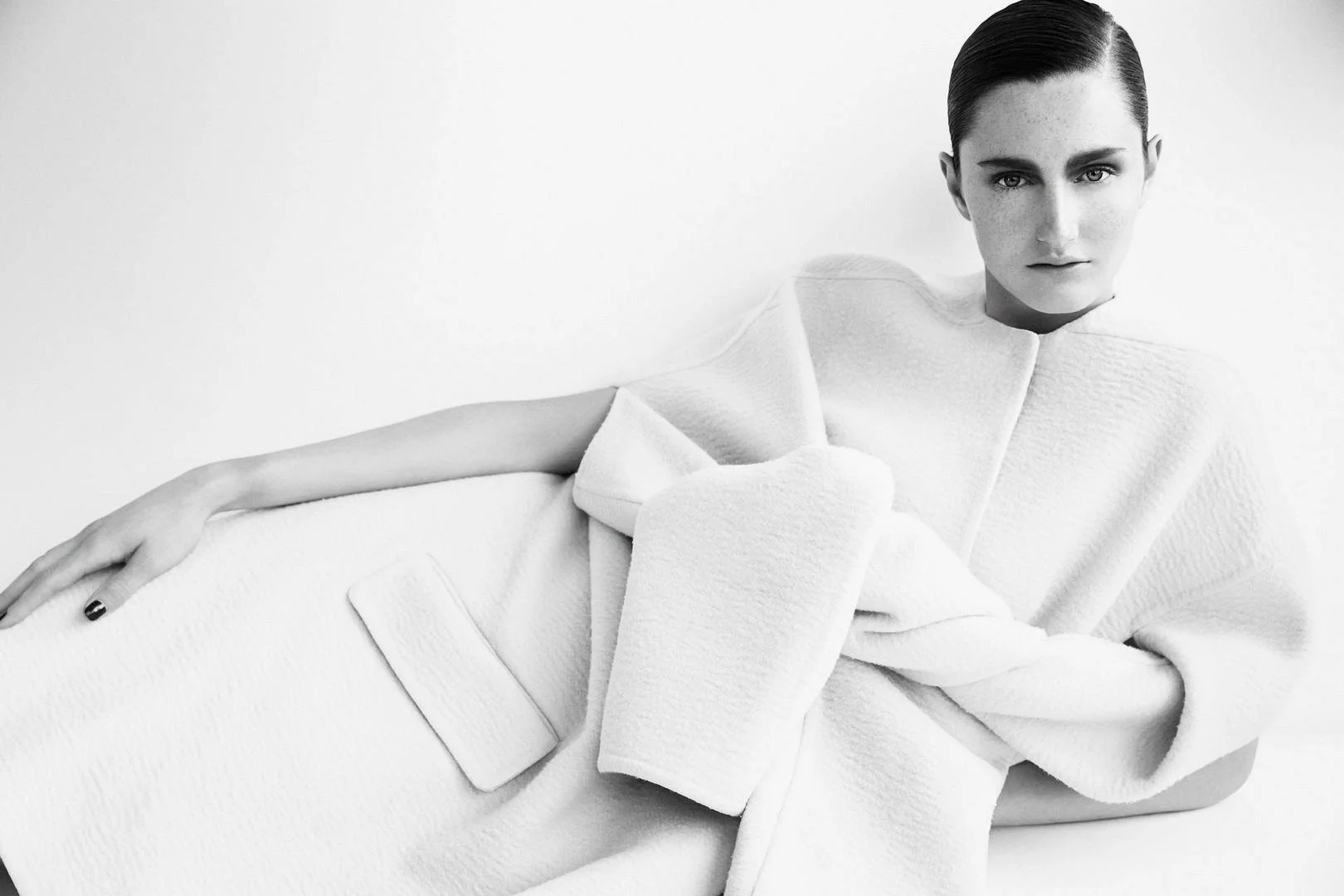 Black and white portrait of a woman with slicked-back hair wearing a textured, structured dress, lying down with her arm extended, against a plain background.