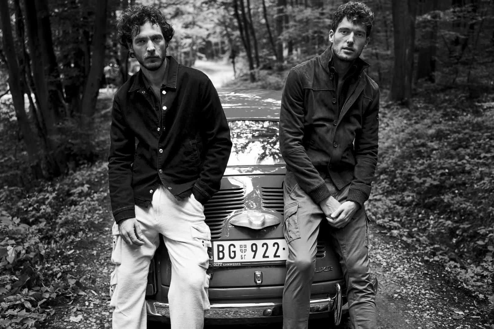 Two young men leaning against the hood of a vintage car on a forest road, with trees and foliage in the background, in black and white.