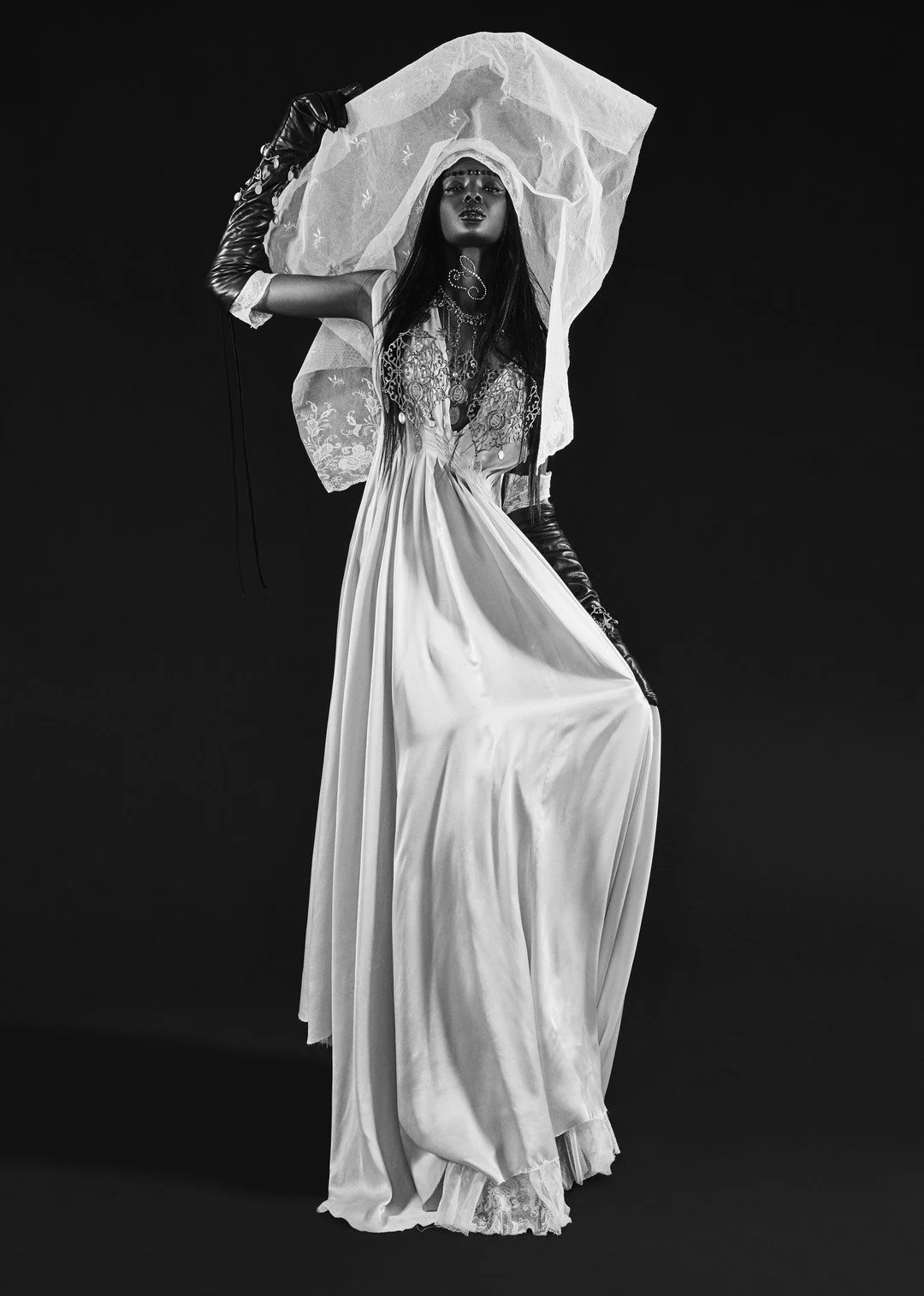 A black and white photograph of a woman dressed in an elegant, vintage-style gown with intricate embroidery and lace details. She wears long gloves, jewelry, and a large veil, striking a dramatic pose against a plain dark background.