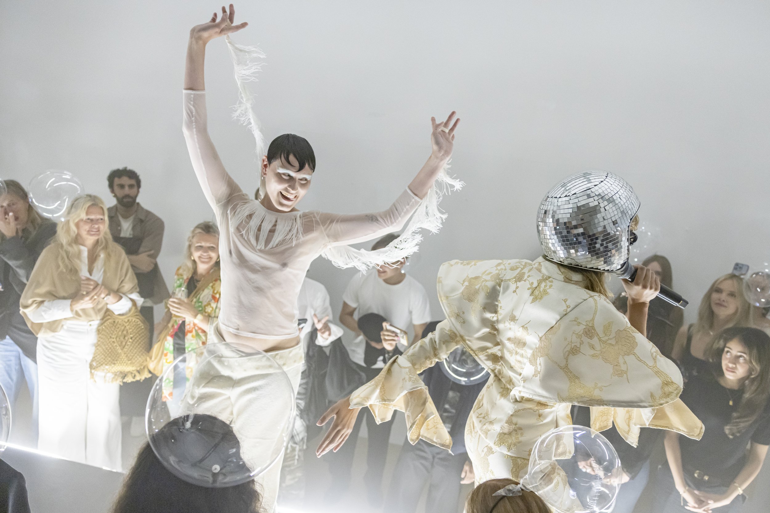 A performer wearing a shiny silver disco helmet and a gold-patterned outfit sings into a microphone on stage. Behind, an audience watches and claps, with some smiling and taking photos. The scene appears lively and festive, with bubble-like effects f